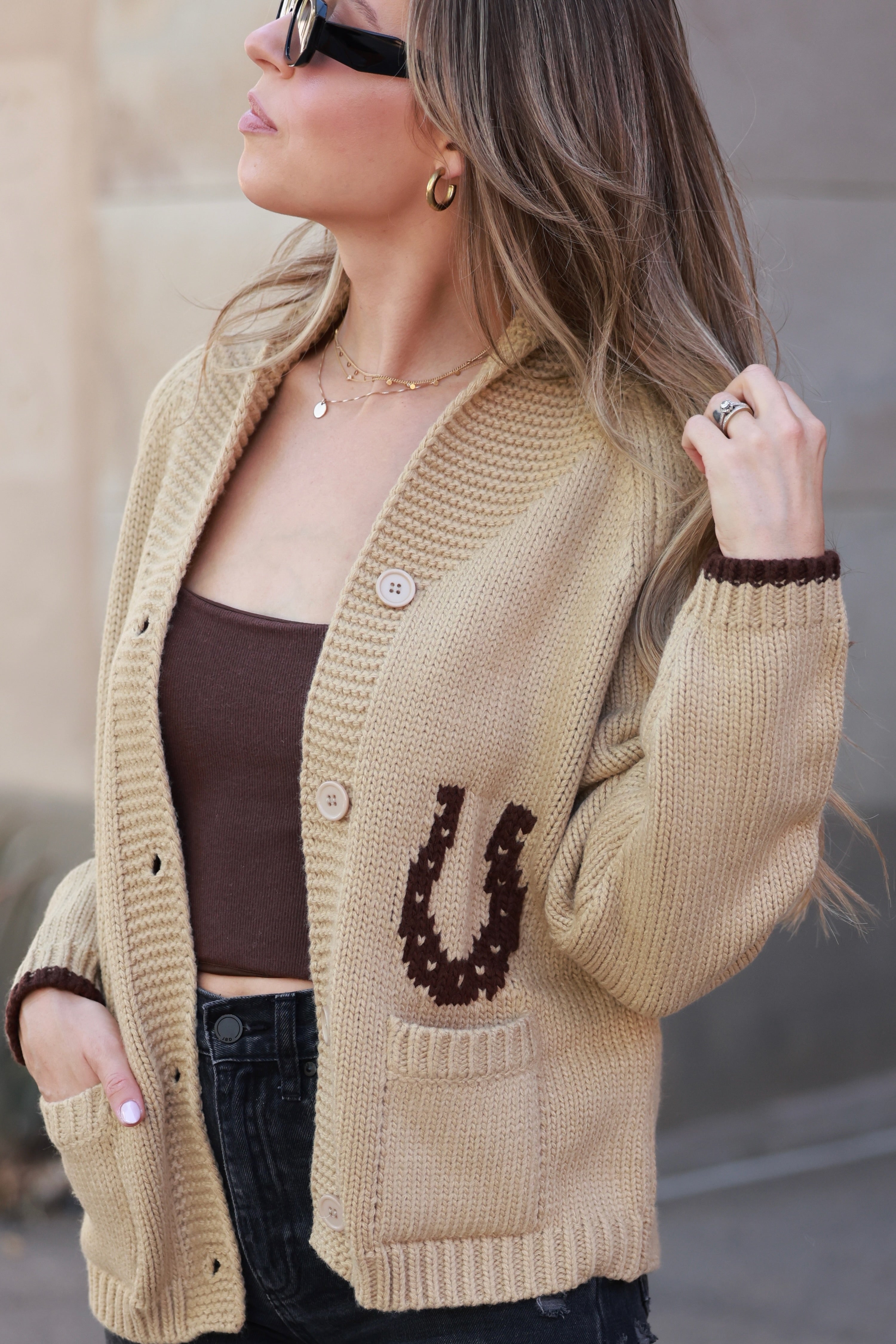 Woman wearing a beige cardigan with a horse shoe design, standing outdoors. - Varsity cardigan, Horse graphic sweater, Equestrian knitwear, Western statement cardigan, Camel horse cardigan