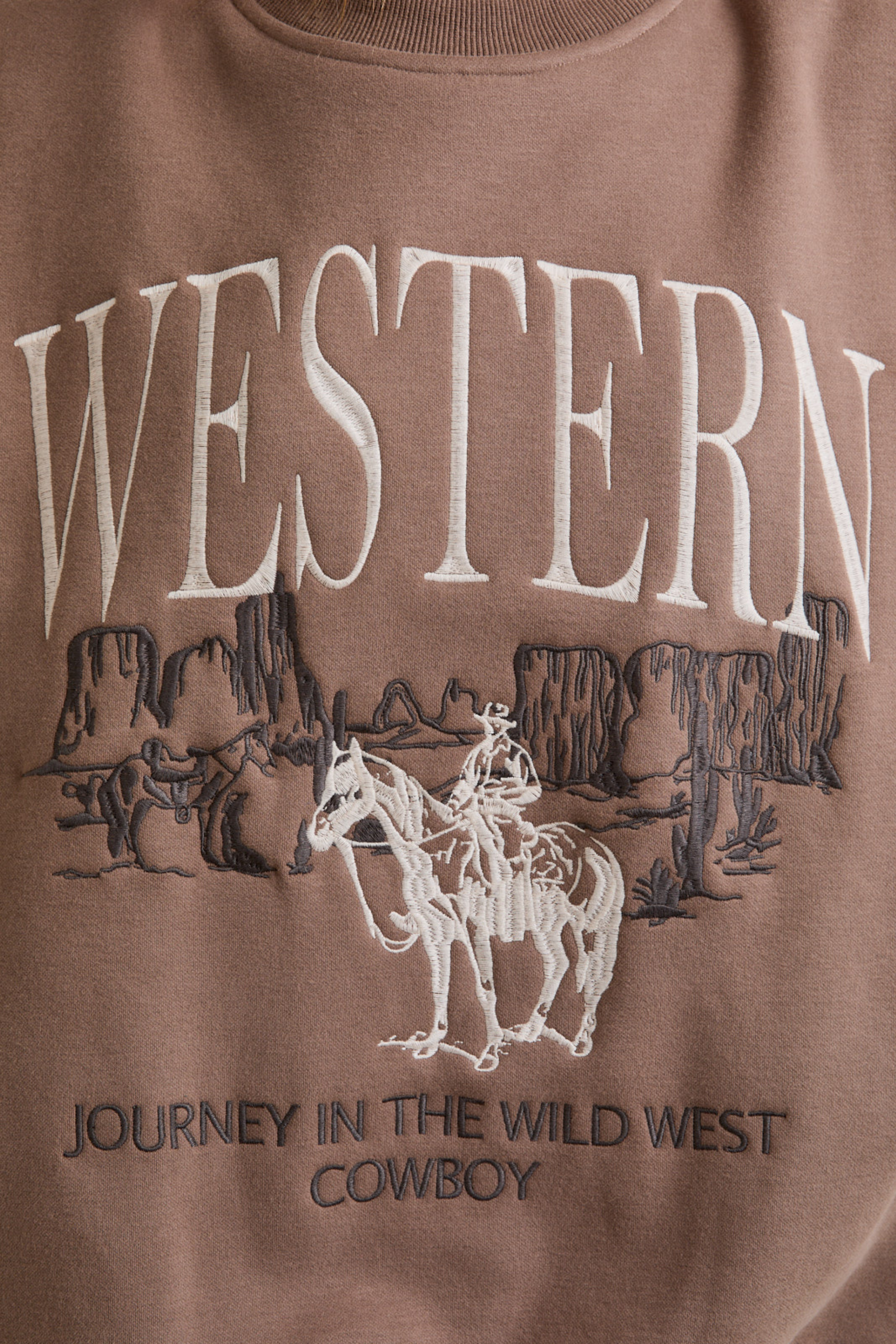 Brown sweatshirt with "WESTERN" in bold white letters, featuring an embroidered cowboy scene with cliffs. Text below reads, "JOURNEY IN THE WILD WEST COWBOY." western graphic sweatshirt, cowboy embroidered sweatshirt, women’s western fashion, vintage cowboy crewneck, casual western outfit, adventure inspired sweatshirt, bold western text sweater, wild west cowboy top, rustic western streetwear