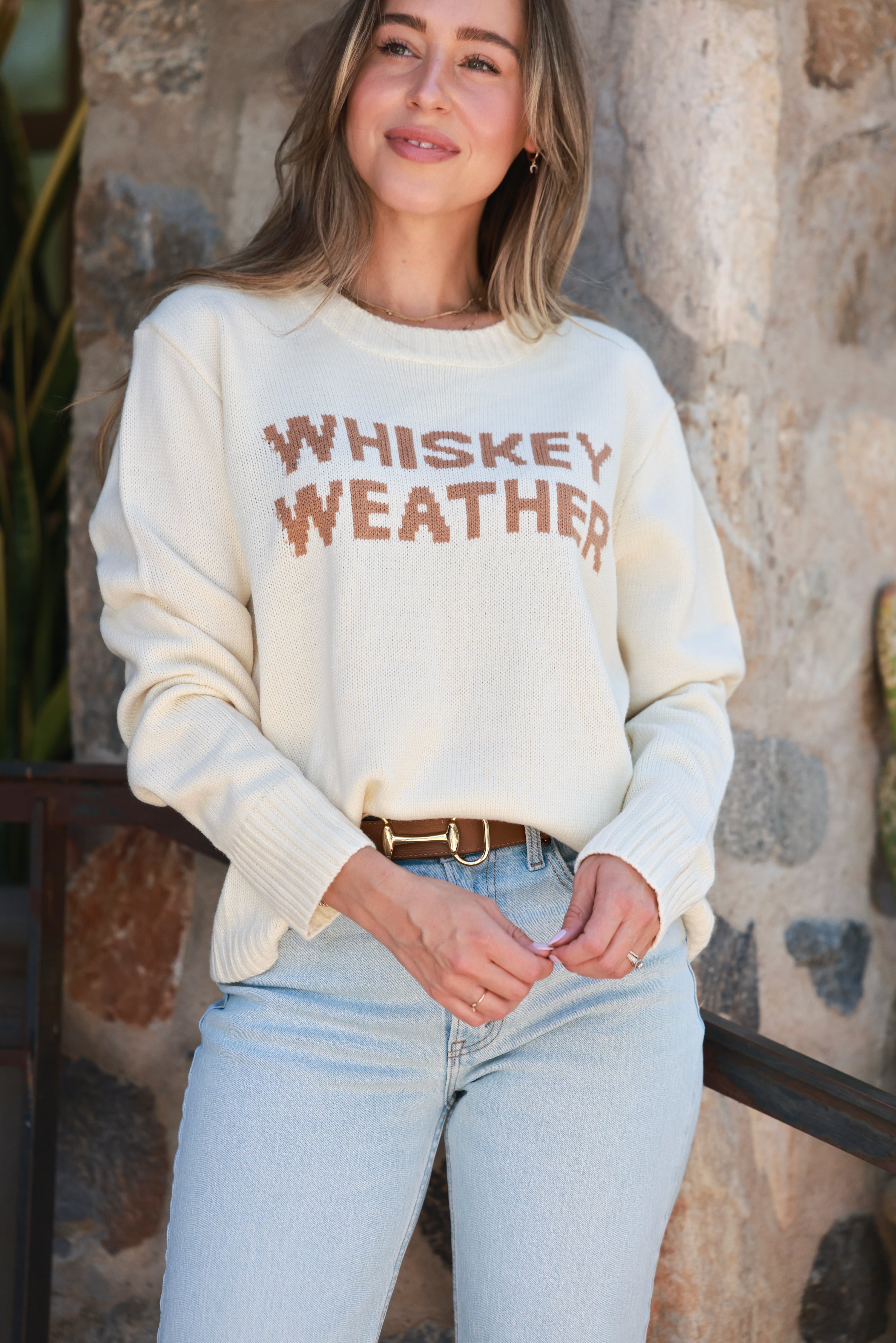 Woman wearing a 'Whiskey Weather' sweater against a stone wall.