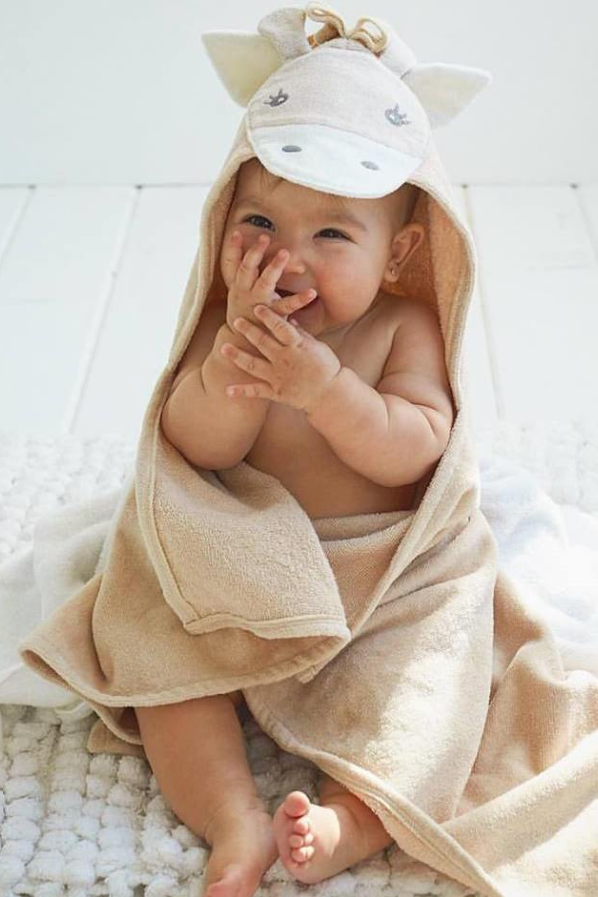 Soft beige hooded baby towel rolled and styled to look like a whimsical pony, featuring a stitched cartoon face, small fabric ears, and a fluffy mane. The towel is tied with a neat white ribbon, highlighting its cozy, giftable presentation. Ideal for newborns to 24 months, the 100% cotton velour terry fabric appears plush and soft, perfect for keeping little ones warm and dry after bath time.