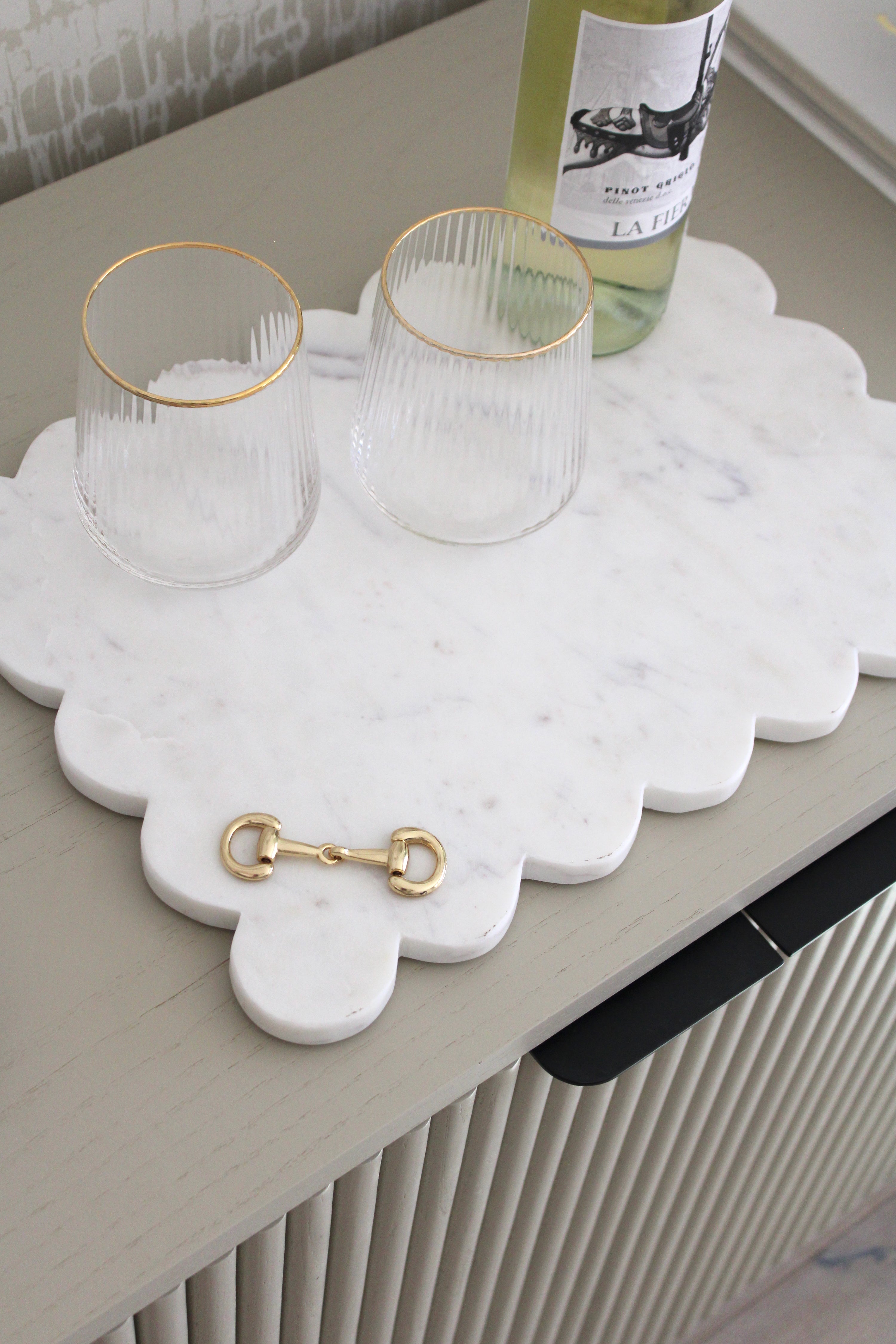 Elegant white marble scalloped serving board with gold D-ring bit, styled in an equestrian-themed kitchen. The handcrafted marble board features subtle natural veining and polished gold hardware, displayed with cheese, fruits, and wine accents. Perfect as a horse-inspired charcuterie tray or decorative equestrian home accent.