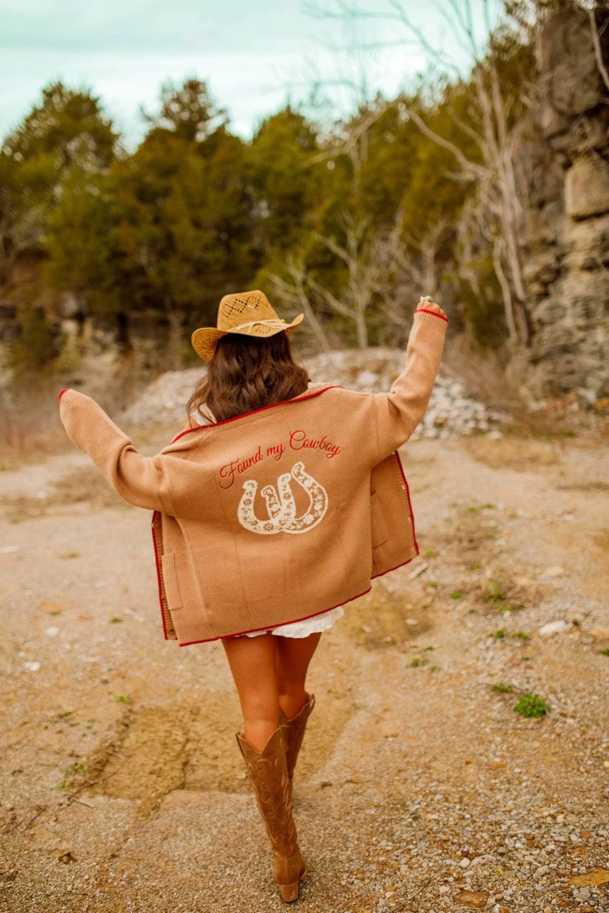 Woman wearing a tan jacket with text and a horseshoe design, standing outdoors with trees and rocks in the background. - Western cardigan, Floral horseshoe sweater, Found my cowboy cardigan, Equestrian-inspired knitwear, Camel embroidered cardigan