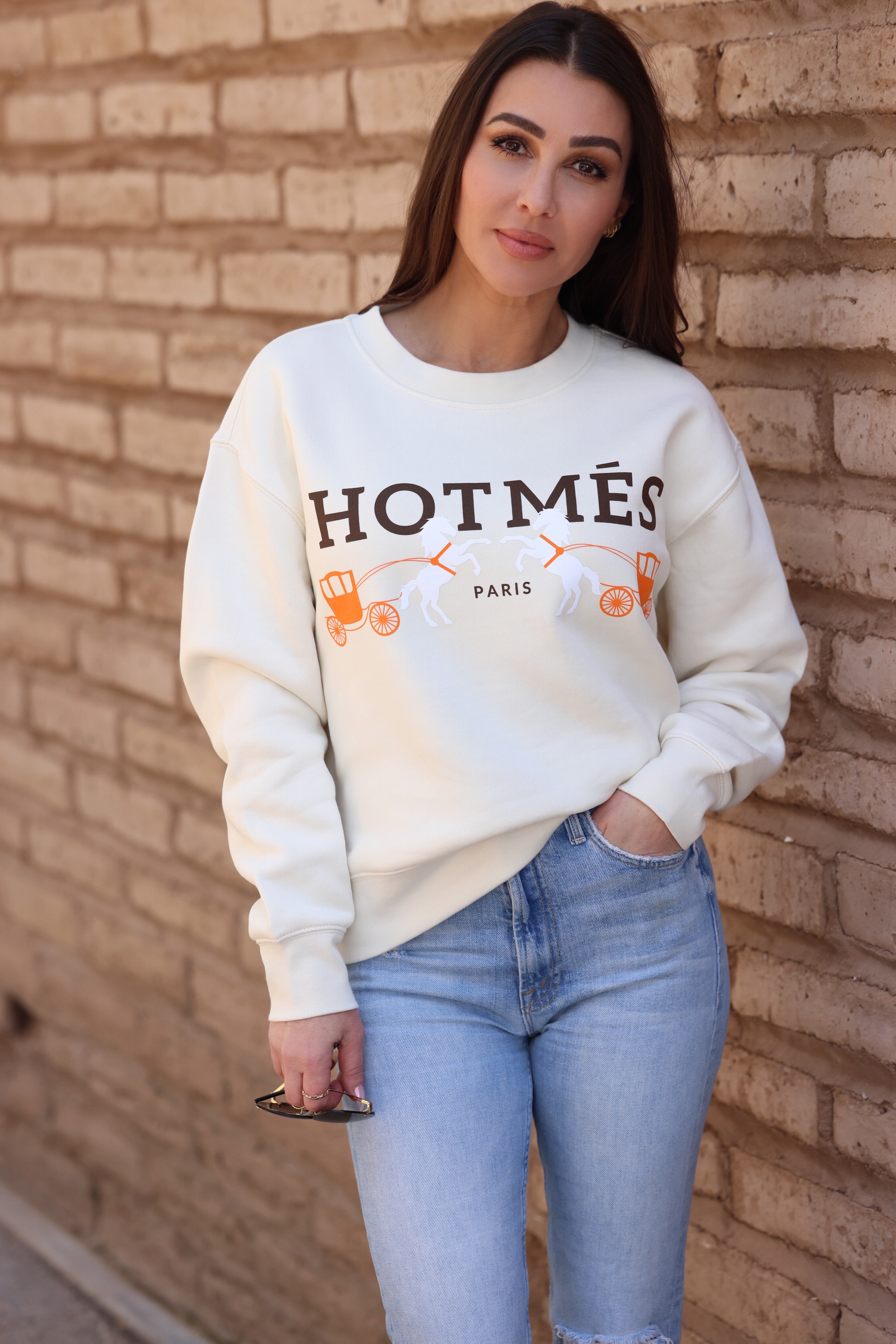 Woman in a white sweatshirt with "HOTMÈS Paris" text and orange graphics, leaning against a brick wall. She holds sunglasses casually, creating a relaxed vibe.