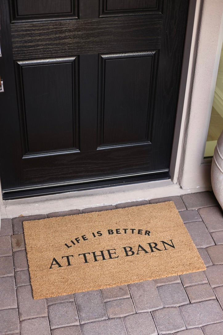 Doormat with "Life is better at the barn" text lies on brick porch.
