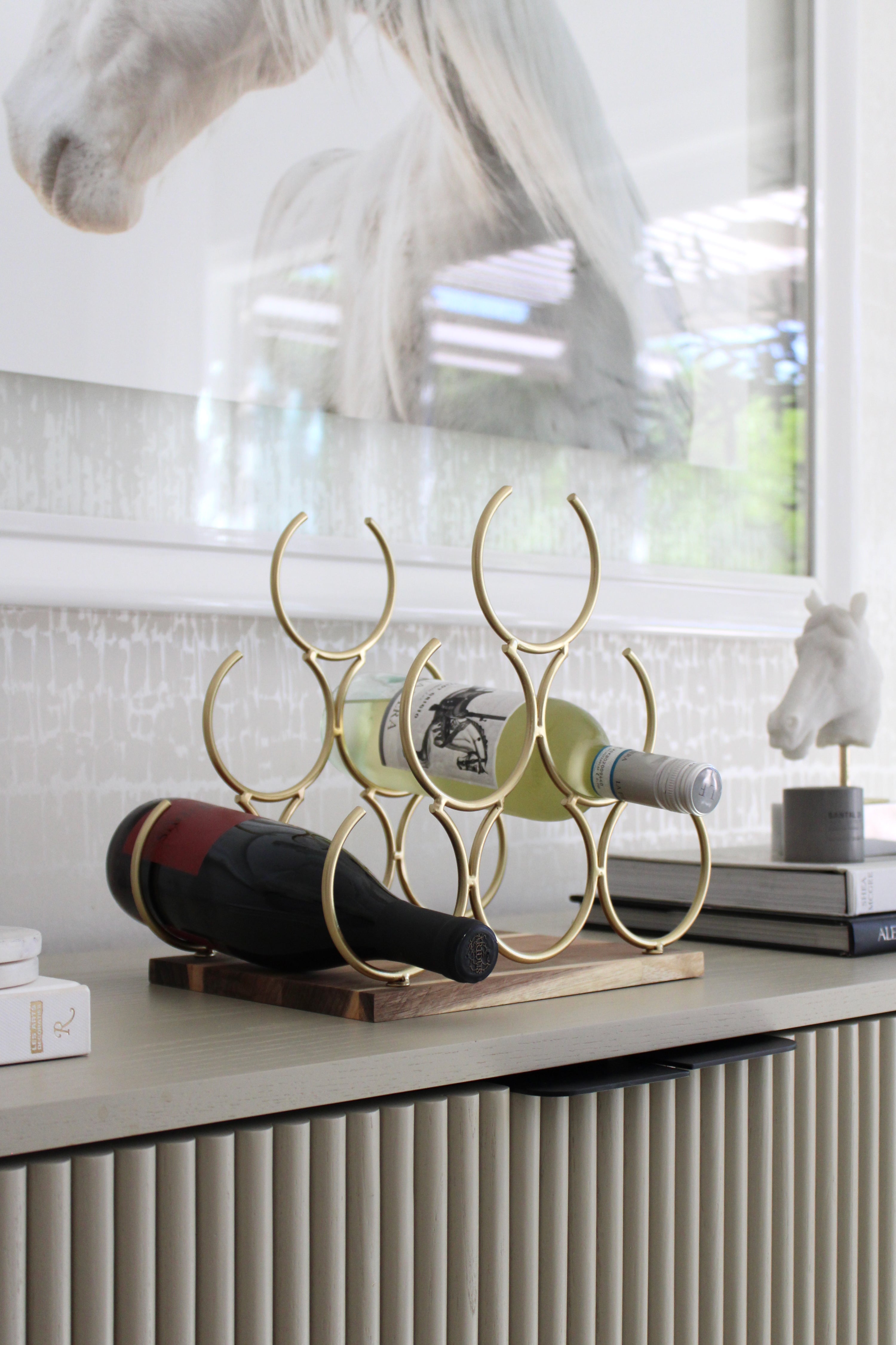 Elegant pyramid-style wine rack with six interlocking gold rings on an acacia wood base, designed to hold up to six wine bottles. The sculptural cast-iron frame with a polished gold finish adds a luxurious equestrian-inspired touch to kitchen or bar décor.