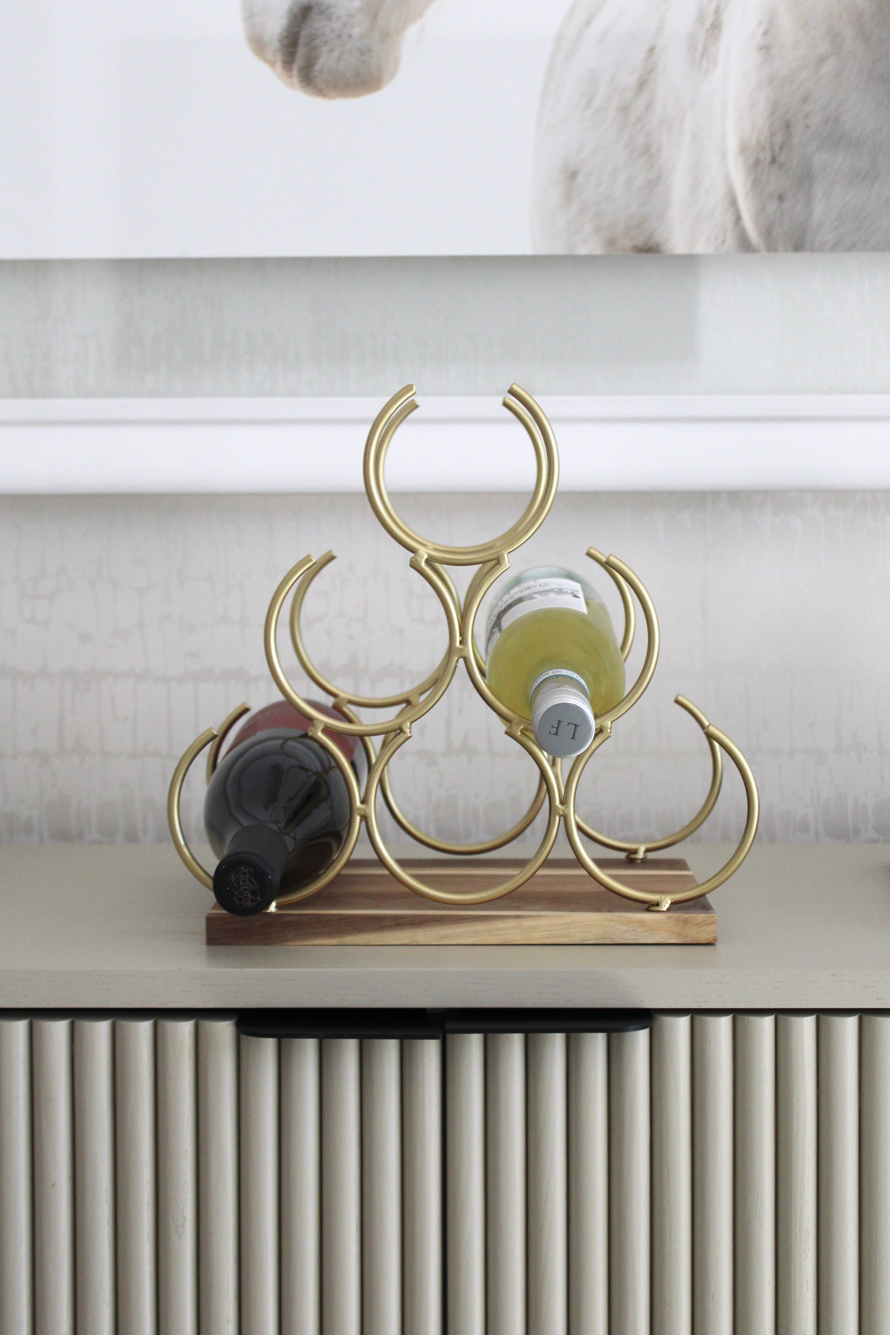 Elegant pyramid-style wine rack with six interlocking gold rings on an acacia wood base, designed to hold up to six wine bottles. The sculptural cast-iron frame with a polished gold finish adds a luxurious equestrian-inspired touch to kitchen or bar décor.