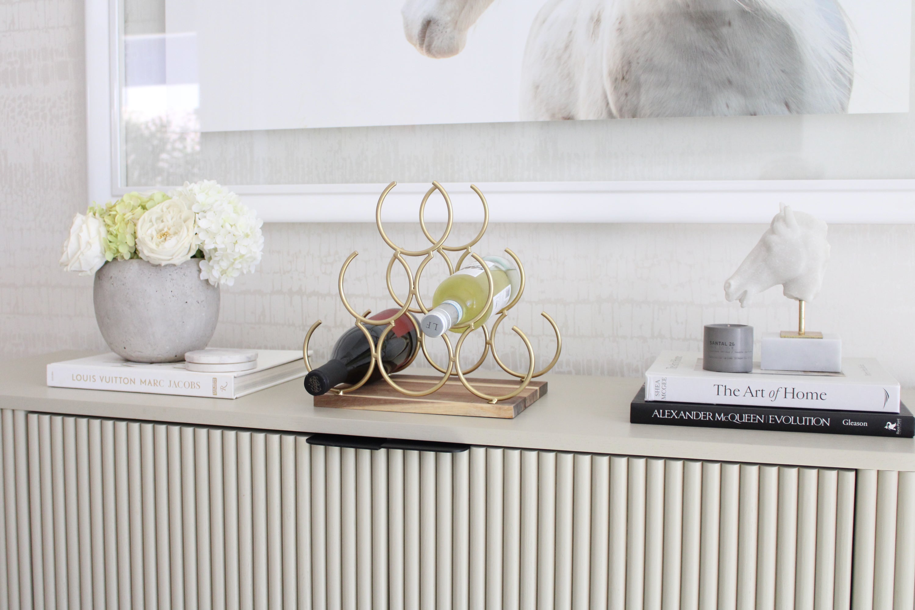 Elegant pyramid-style wine rack with six interlocking gold rings on an acacia wood base, designed to hold up to six wine bottles. The sculptural cast-iron frame with a polished gold finish adds a luxurious equestrian-inspired touch to kitchen or bar décor.