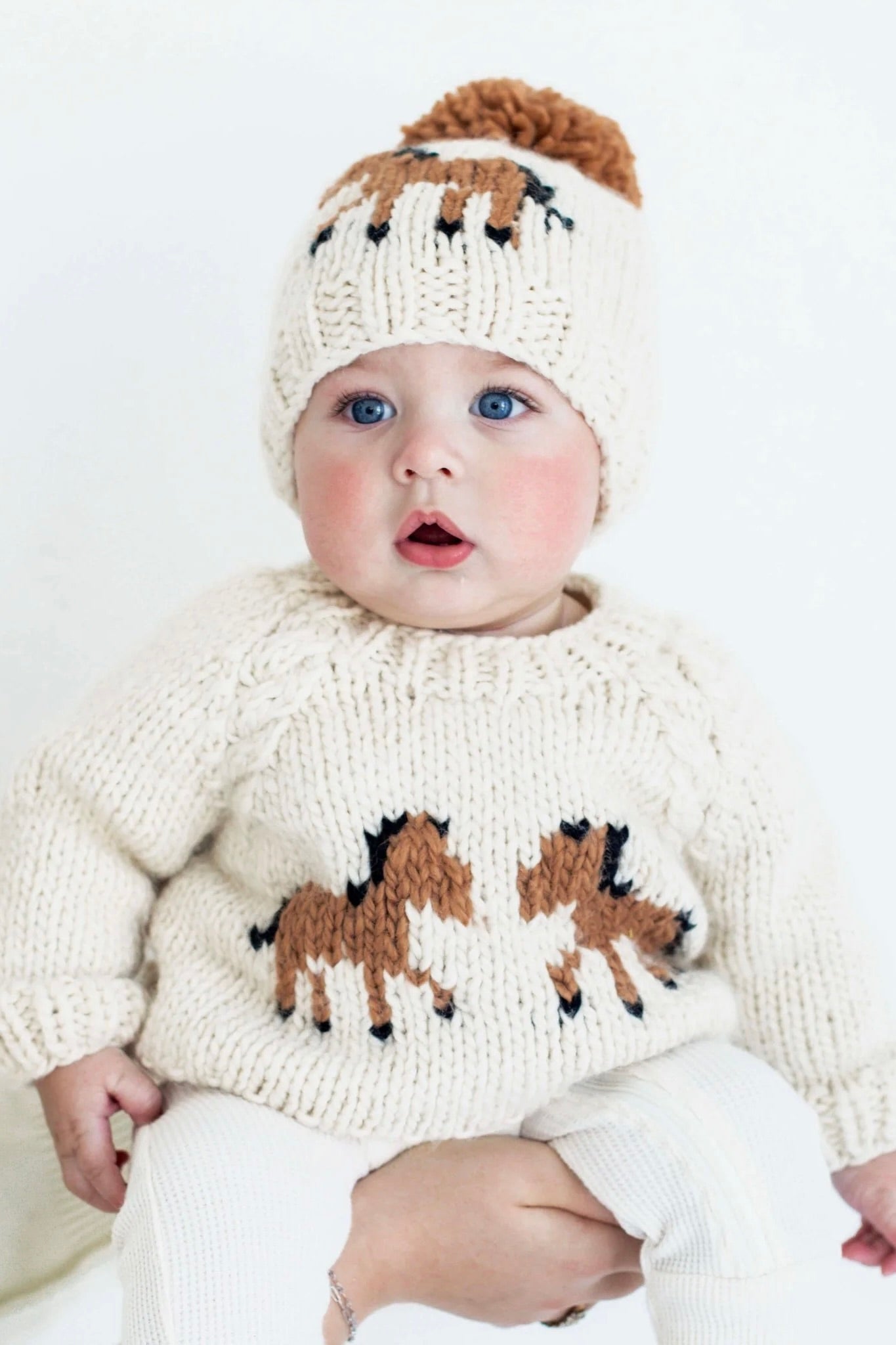 Baby wearing a cream, hand-knit pony sweater made of soft, sustainable yarn with two brown horses facing each other on the front, raglan sleeves, and cable accents, paired with a matching pom-pom beanie. Cozy, playful design conveys warmth and charm, photographed against a plain background.