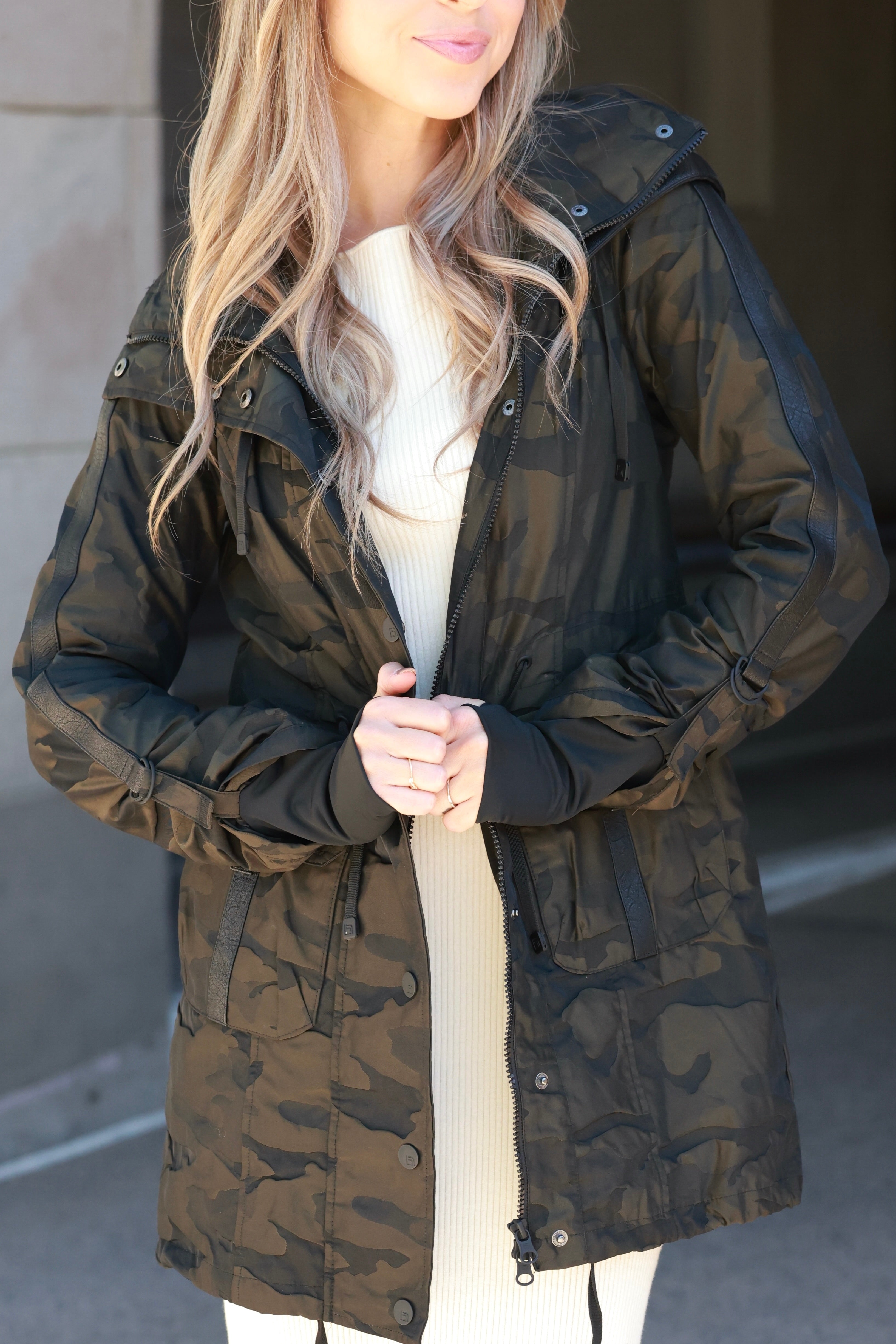 Woman wearing an olive camo anorak jacket with adjustable waist, hood, storm cuffs with thumbholes, zip front with button closure, and two front pockets. Stylish equestrian outerwear designed as a versatile horse riding jacket, barn jacket, and raincoat for horse girls.