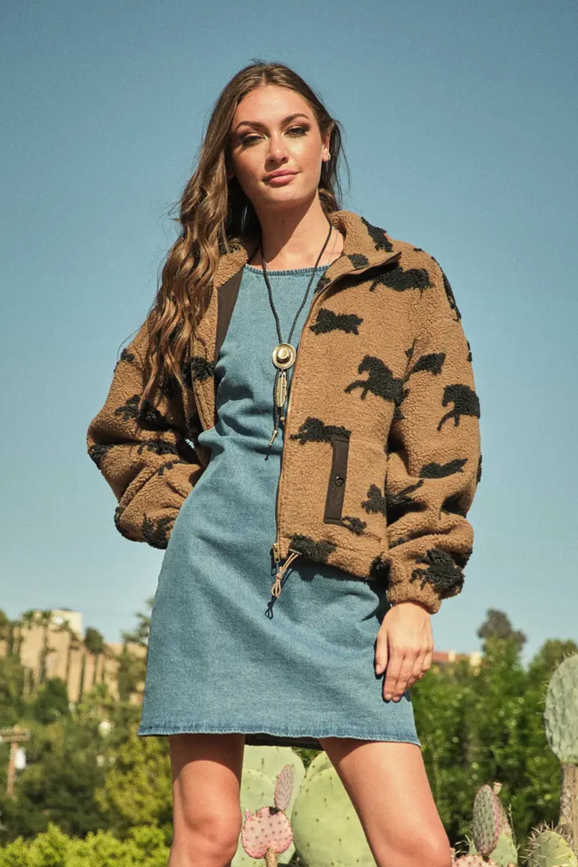 Woman wearing a brown horse patterned jacket over a denim dress, stands outdoors against a clear blue sky, with lush greenery and cacti in the background.