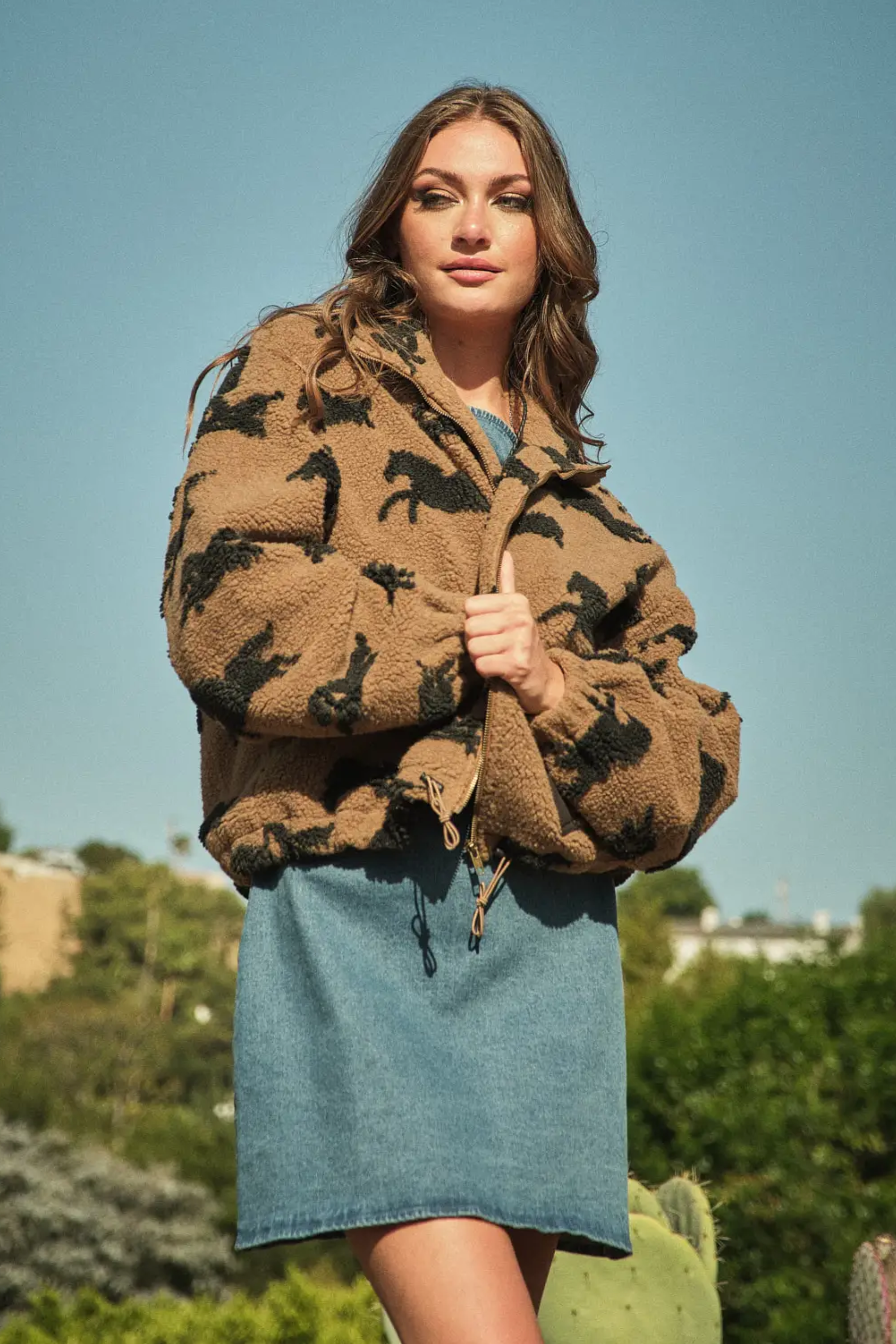 Woman wearing a brown horse patterned jacket over a denim dress, stands outdoors against a clear blue sky, with lush greenery and cacti in the background.