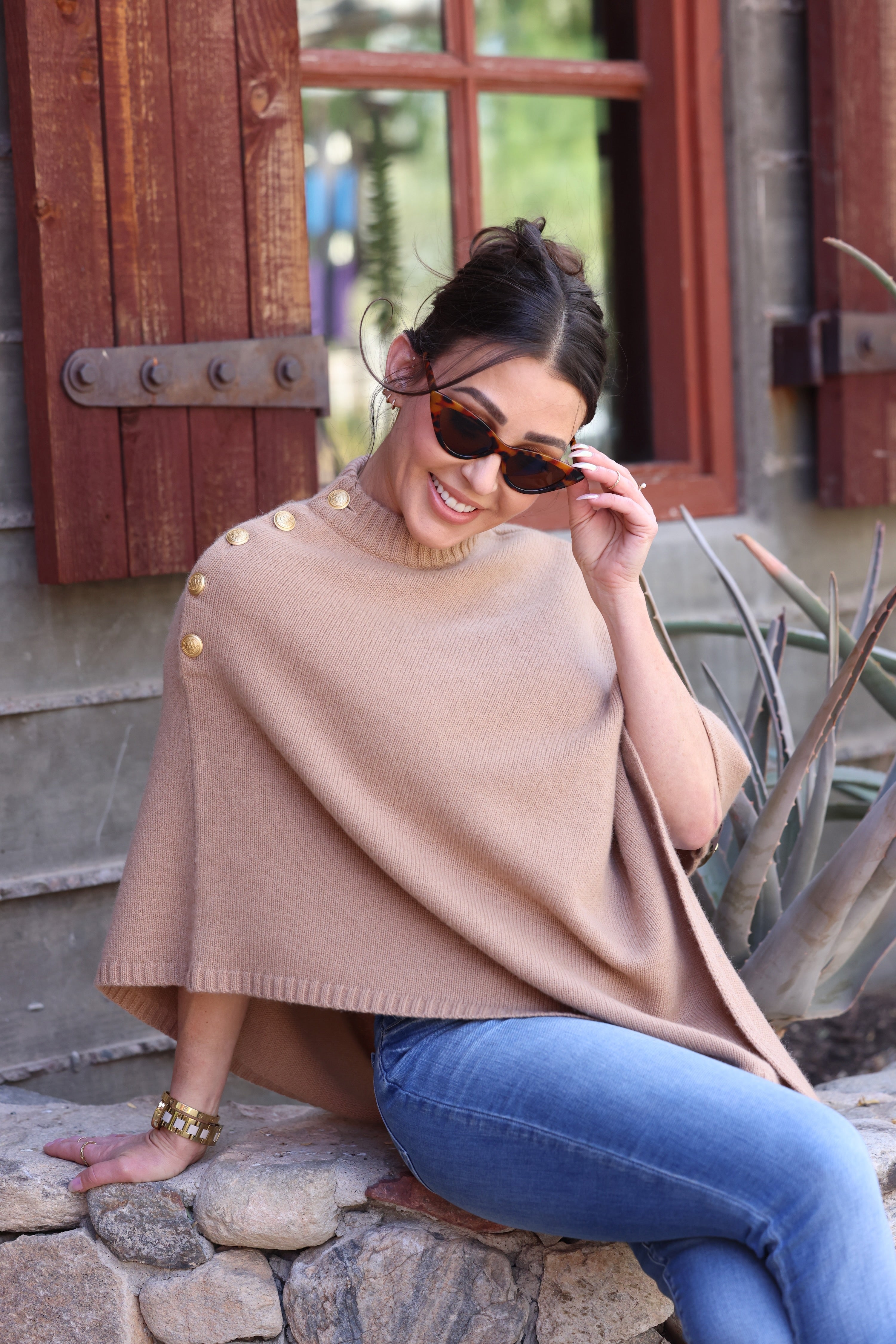 Smiling woman in sunglasses and camel colored poncho with gold buttons sits on stone bench, surrounded by rustic wooden window shutters and plants.