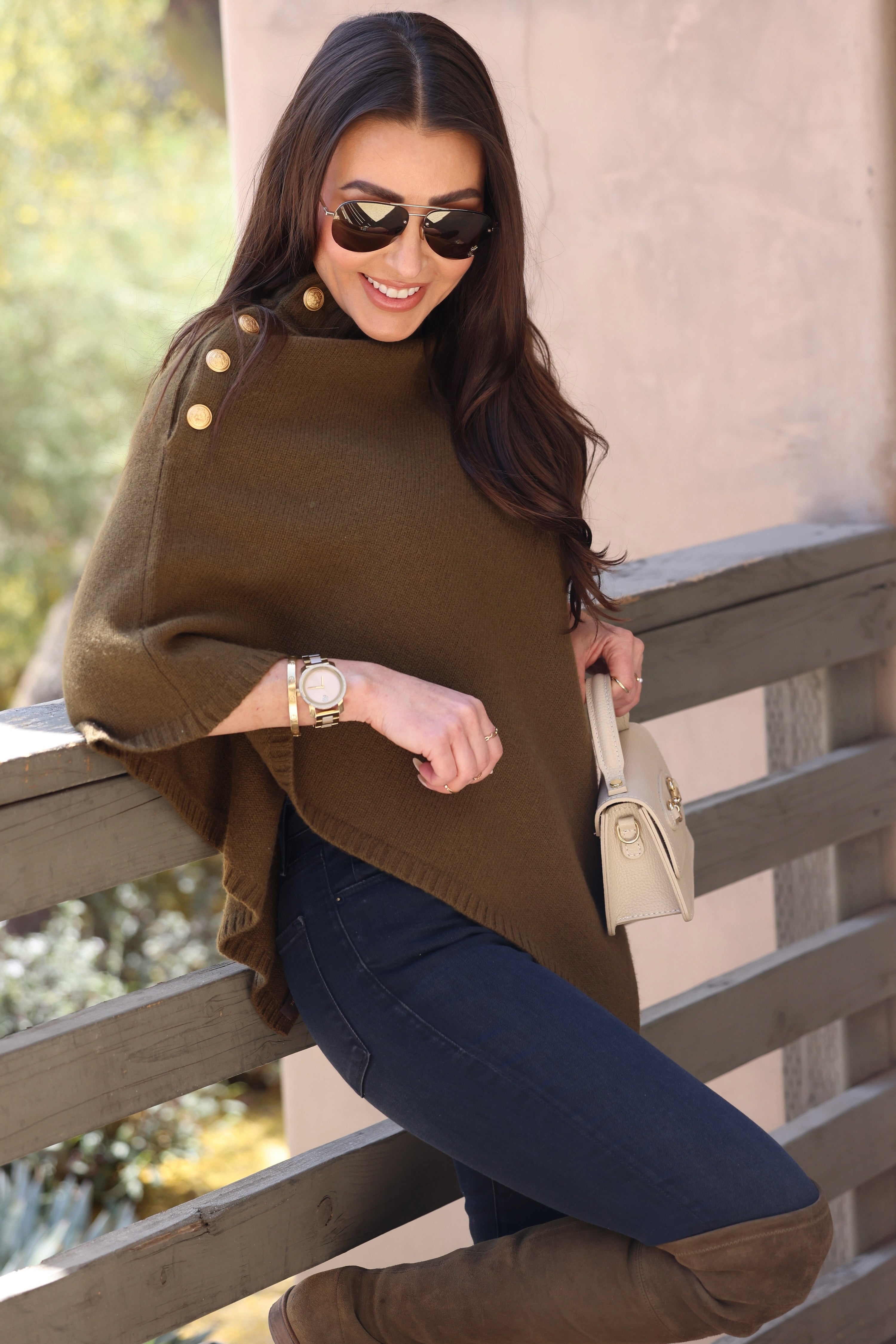 Woman in a olive poncho with gold buttons, wearing aviator sunglasses and a gold watch, leans on a wooden railing outdoors, smiling, holding a beige handbag.