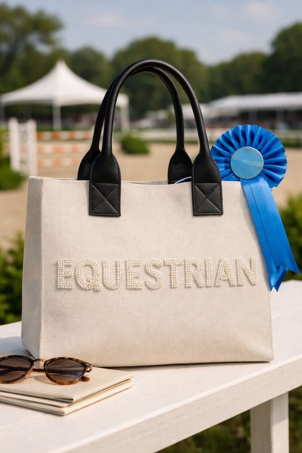 Beige tote bag with 'Equestrian' text, black handles, and a blue ribbon on a white surface outdoors.