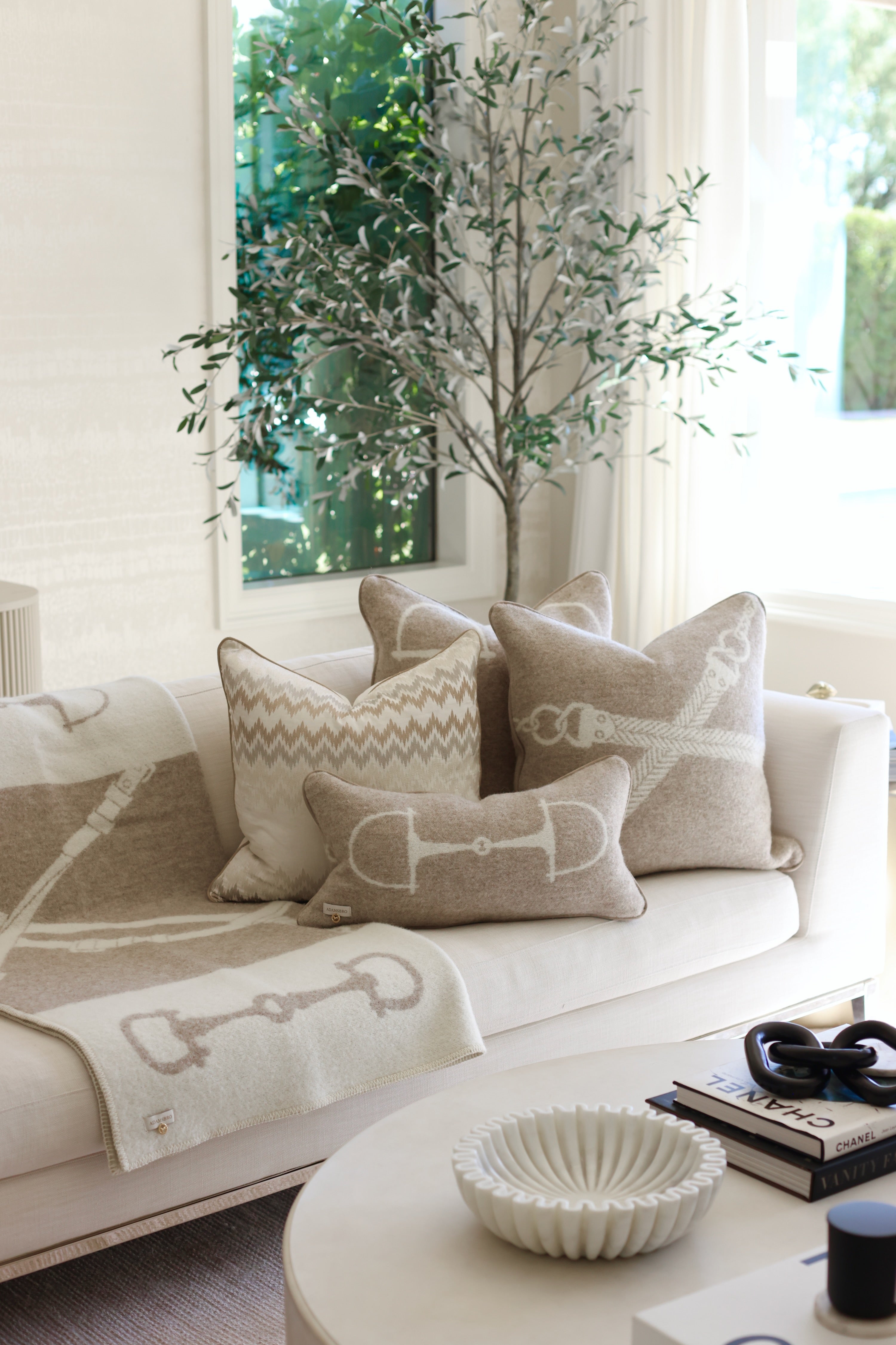 Equine Lumbar Wool Pillow - Vanilla Bean A cozy living room scene with a white sofa adorned with beige and white patterned pillows and a matching blanket. A green indoor tree stands beside it. Luxury rectangular cushion in vanilla bean, made from 100% New Zealand lambswool with ADAMSBRO’s signature equestrian bit pattern. Handcrafted in Sweden, featuring faux suede piping, concealed zipper, and a plush duck feather insert, a refined accent for timeless, Scandinavian-inspired interiors.