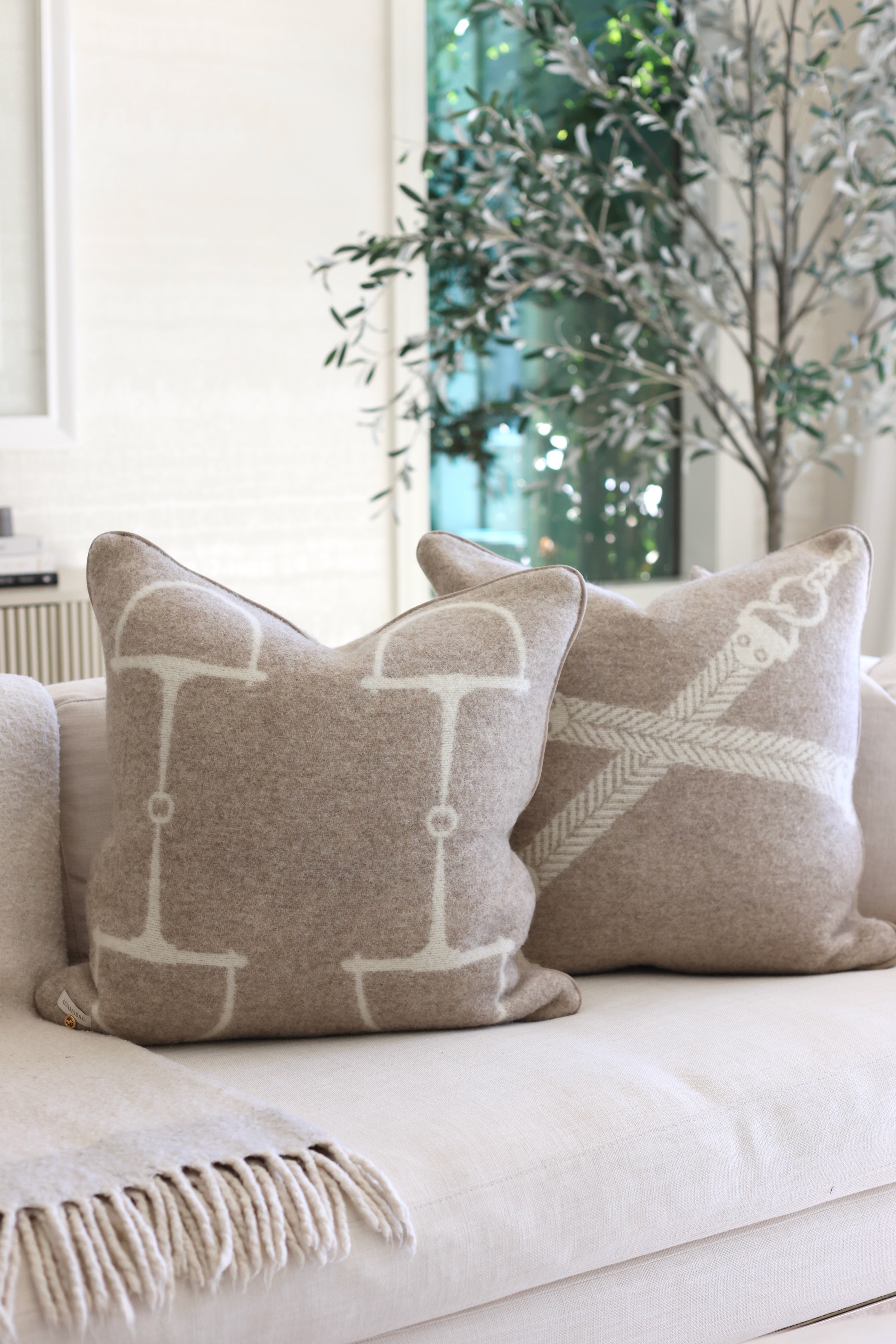 Adamsbro Equine Wool Pillow - Vanilla Bean | Cozy beige sofa with two horse bit patterned pillows; soft texture and neutral tones. A potted plant and framed art create a serene, minimalist ambiance. Luxury cushion in 100 new zealand lambswool | Luxury rectangular cushion handcrafted in Sweden from 100% New Zealand lambswool featuring an equestrian pattern, soft faux suede piping, and filled with plush duck feathers, a Scandinavian-inspired accent pillow
