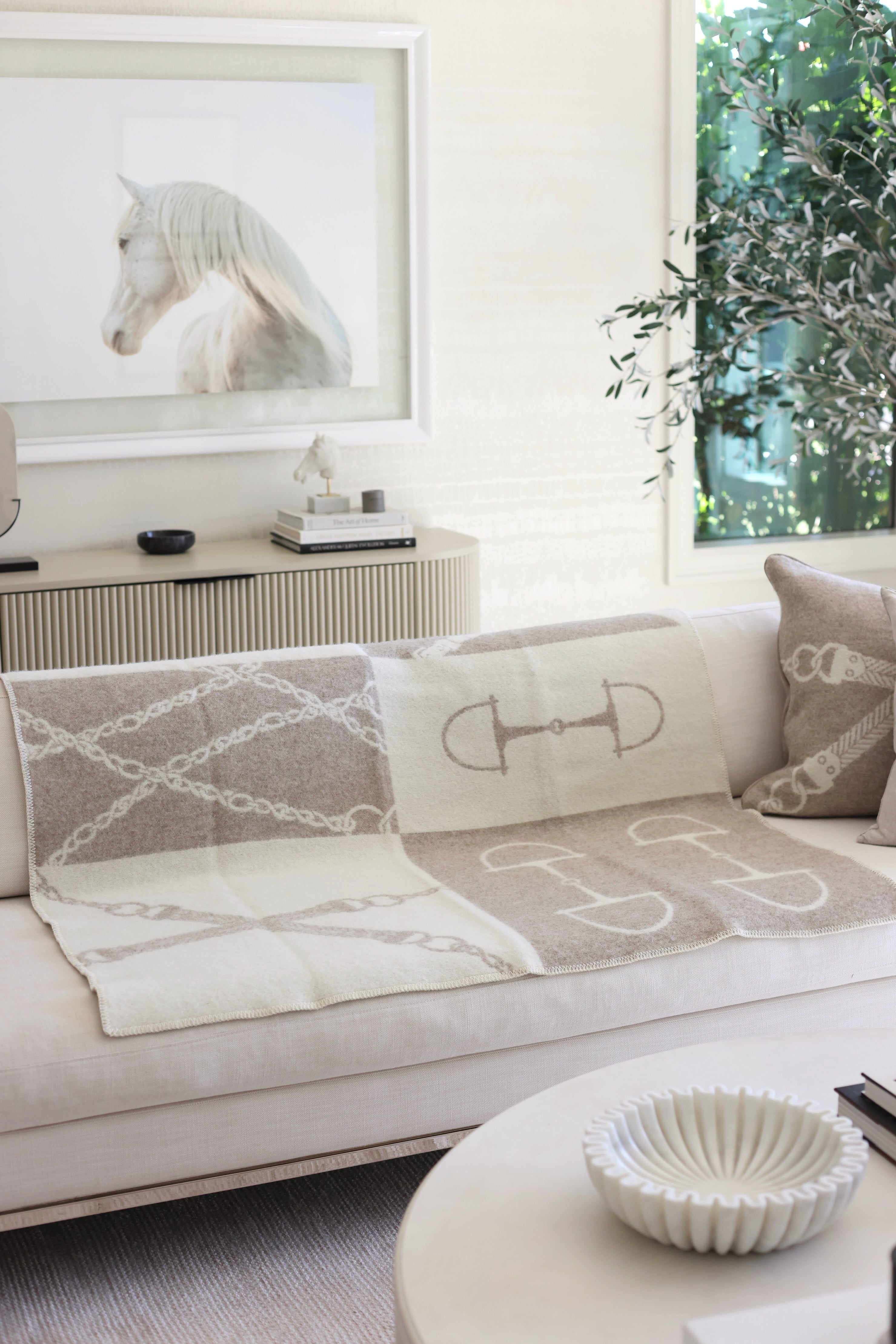 Equine Wool Throw - Vanilla Bean | A cozy living room features a beige sofa with a horse-themed blanket and pillow. A framed horse photo and potted plant add a serene touch. Classic equestrian-inspired wool throw blanket in vanilla bean, crafted from 100% New Zealand lambswool. Features a reversible horse bit pattern, soft and breathable texture, and Scandinavian design, perfect for layering on sofas, beds, or armchairs in timeless, natural interiors