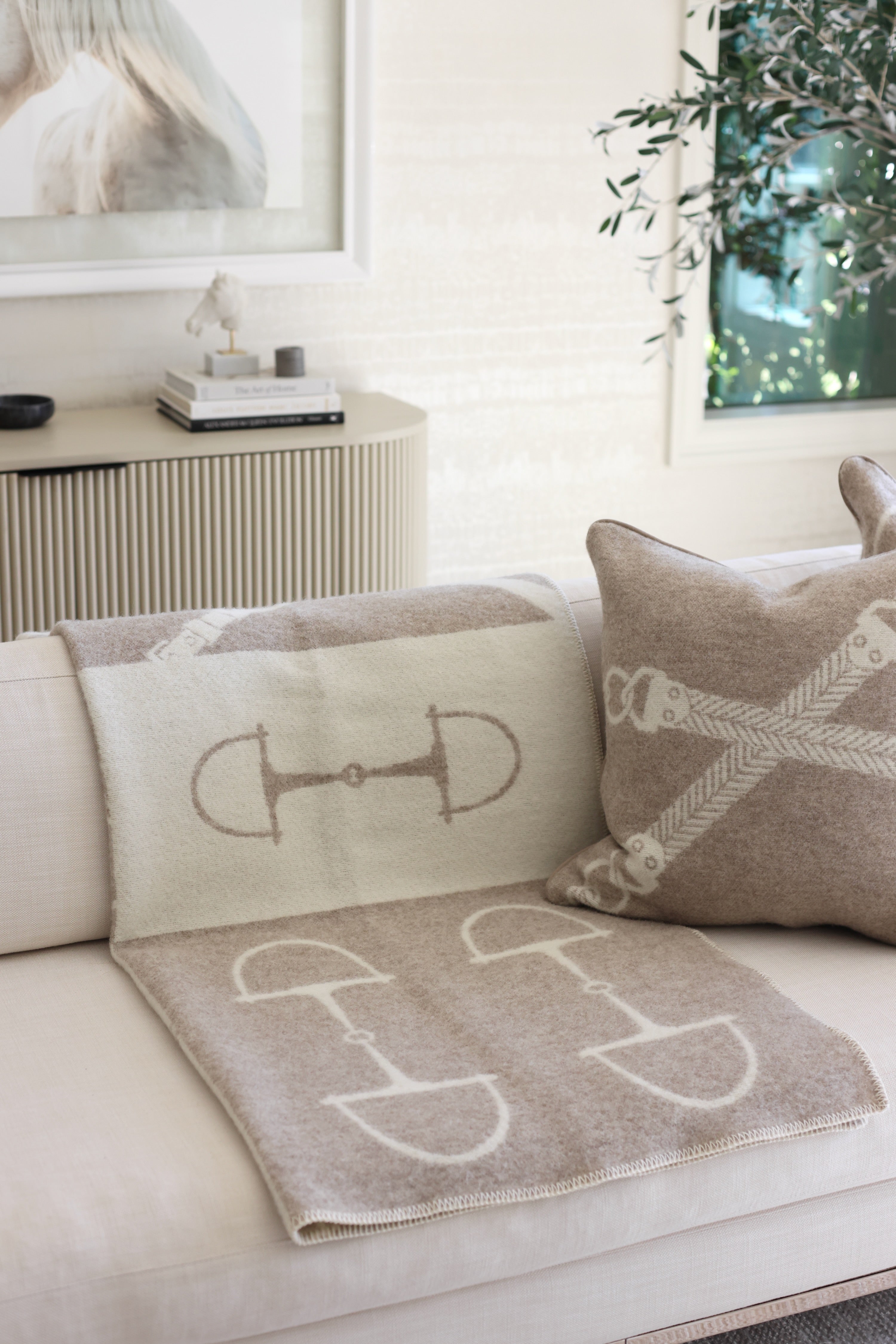 Equine Wool Throw - Vanilla Bean | A cozy living room features a beige sofa with a horse-themed blanket and pillow. A framed horse photo and potted plant add a serene touch. Classic equestrian-inspired wool throw blanket in vanilla bean, crafted from 100% New Zealand lambswool. Features a reversible horse bit pattern, soft and breathable texture, and Scandinavian design, perfect for layering on sofas, beds, or armchairs in timeless, natural interiors