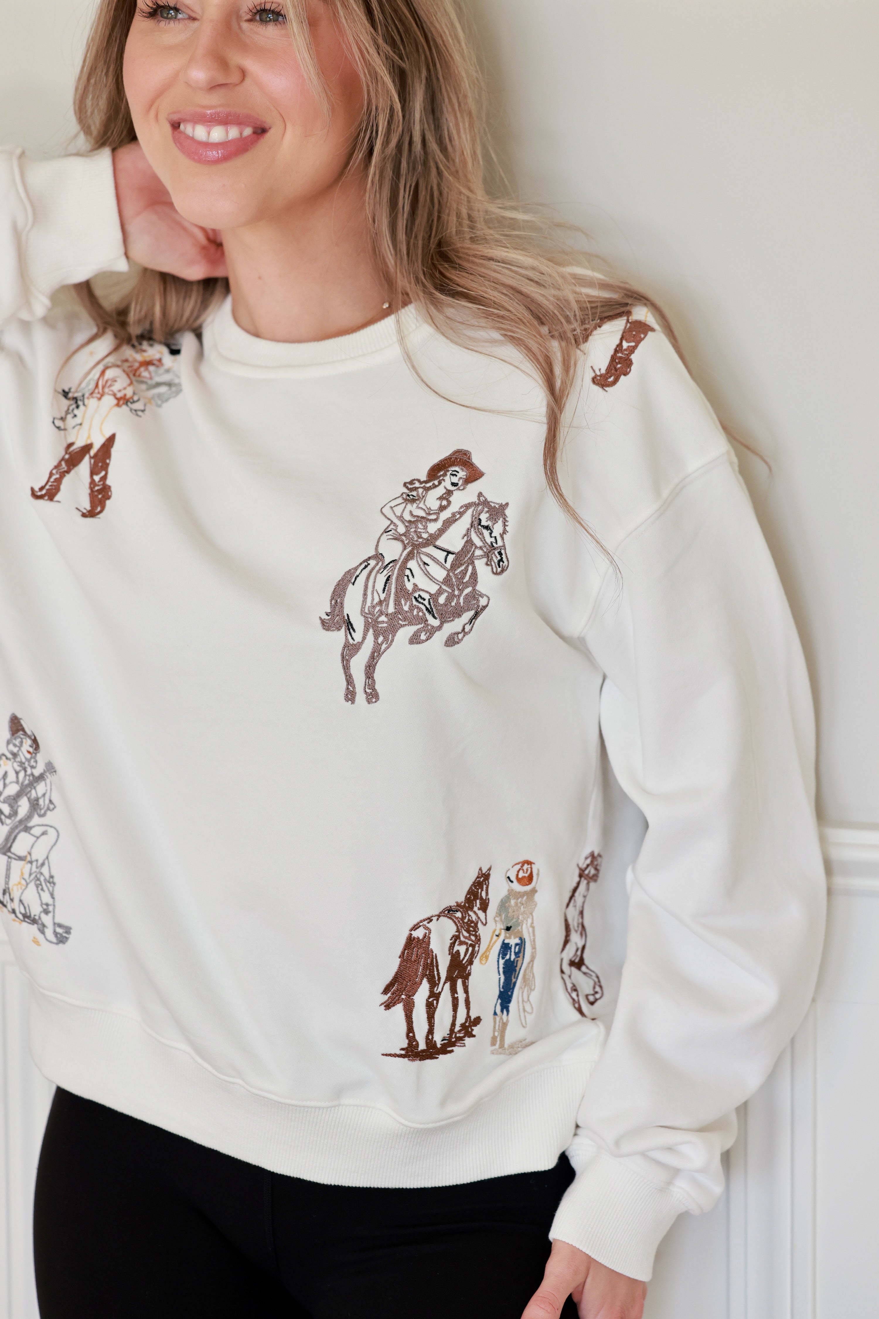 Woman wearing a white embroidered western cowgirl sweatshirt featuring horse and cowgirl-themed illustrations. This cozy equestrian clothing piece blends comfort and western riding style, perfect horse girl apparel with detailed embroidery for everyday wear.