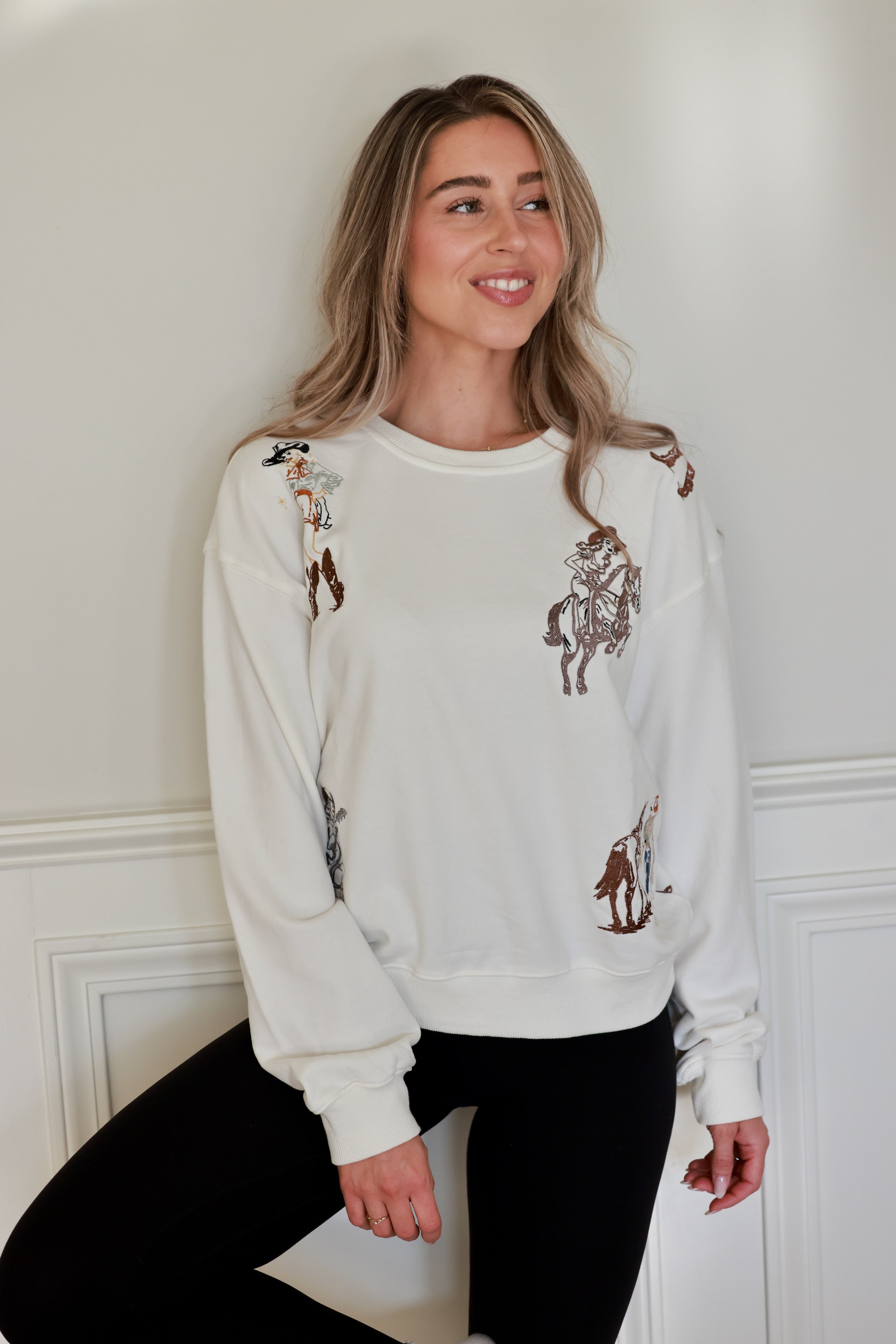 Woman wearing a white embroidered western cowgirl sweatshirt featuring horse and cowgirl-themed illustrations. This cozy equestrian clothing piece blends comfort and western riding style, perfect horse girl apparel with detailed embroidery for everyday wear.