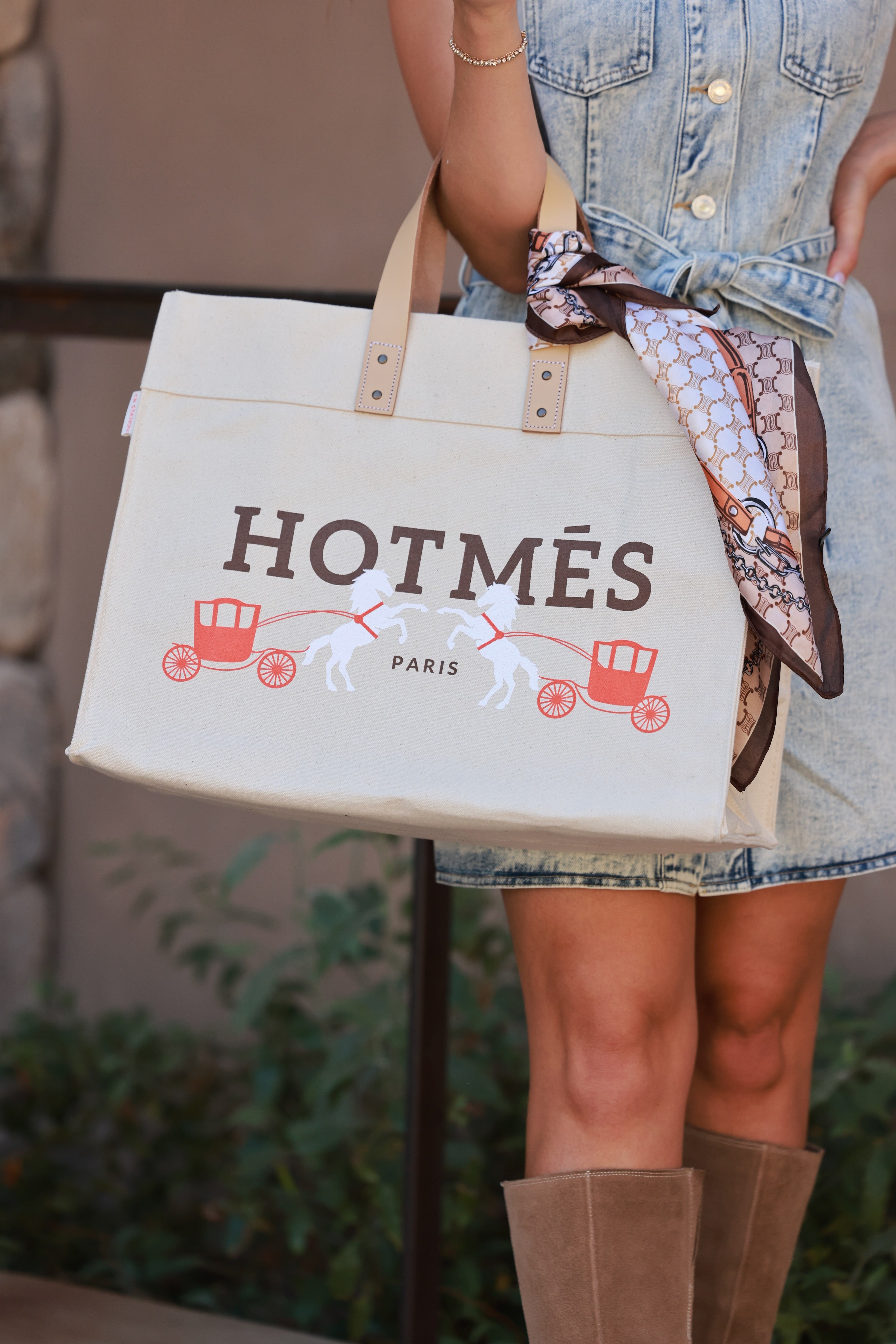 Person holding a 'HOTMéS' bag with a scarf, wearing a denim dress and brown boots. - equestrian accessory, horse themed bag, equestrian travel essentials, designer inspired carriage horse print, equestrian carry-on, weekender bag