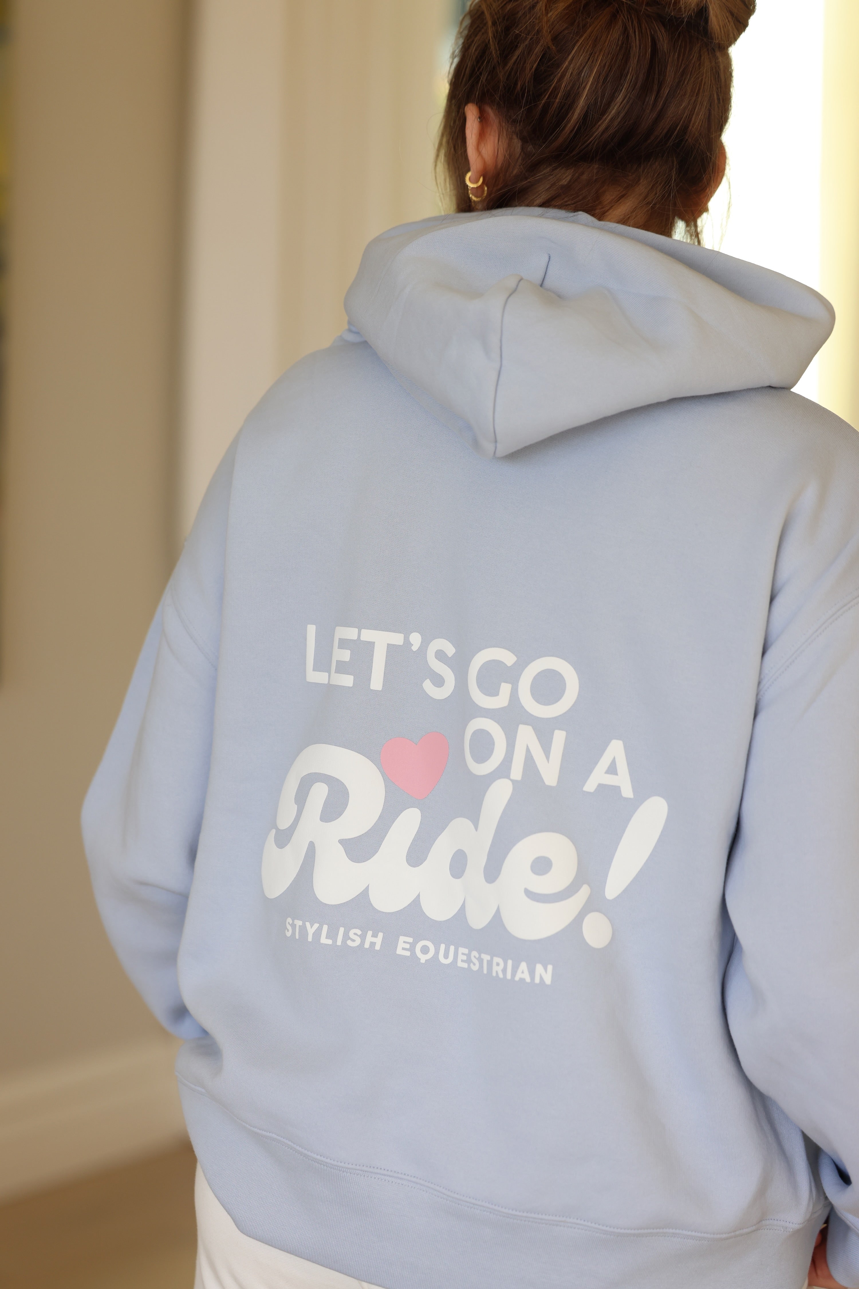 Rear view of a person wearing a light blue hoodie with the text, "Let's Go on a Ride! Stylish Equestrian," and a pink heart. Cozy and inviting tone.