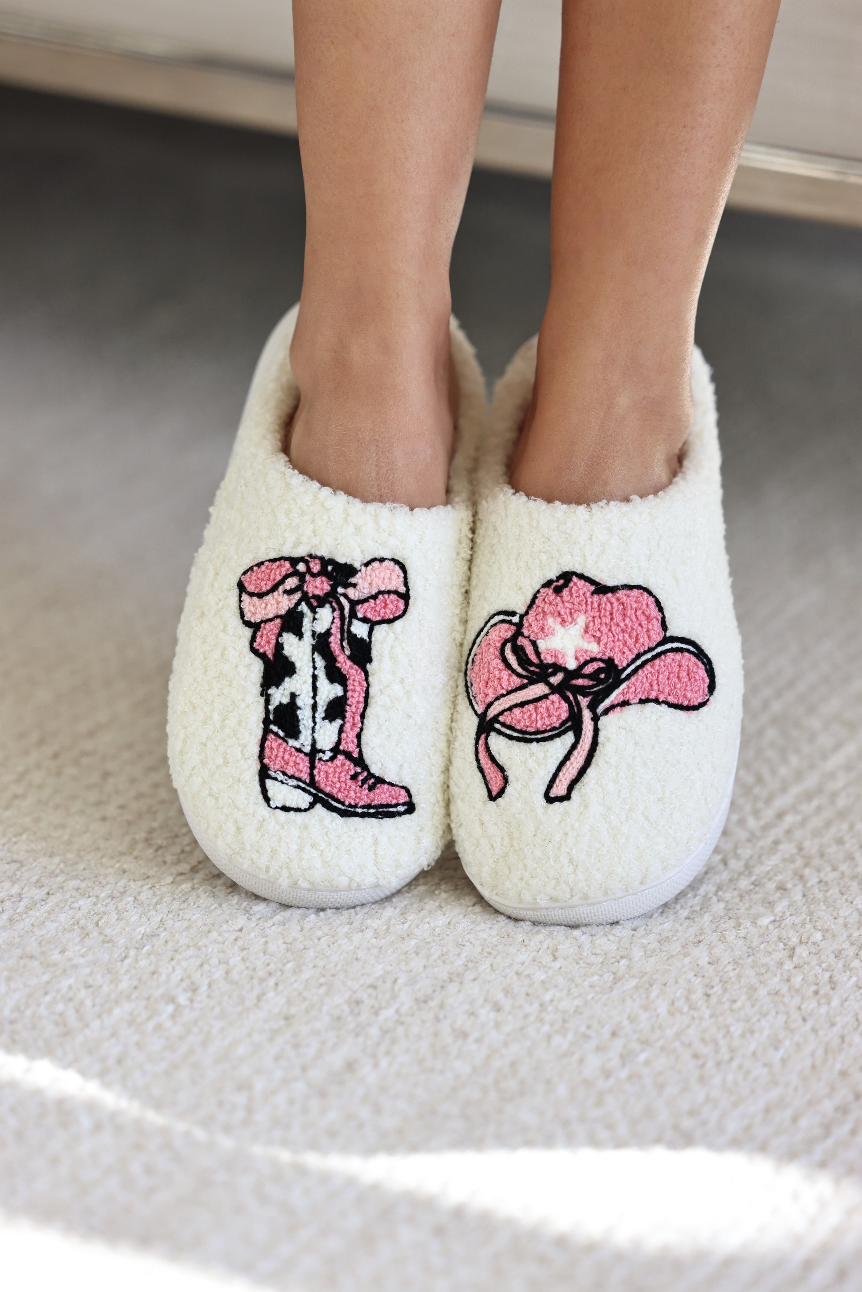 Pink Pony Club Slippers -  A person wearing cozy, off-white fuzzy slippers with playful pink designs—a cowboy boot with a pink bow and cow print on one slipper.  A pink cowboy hat with a white star and pink bow on the other slipper.