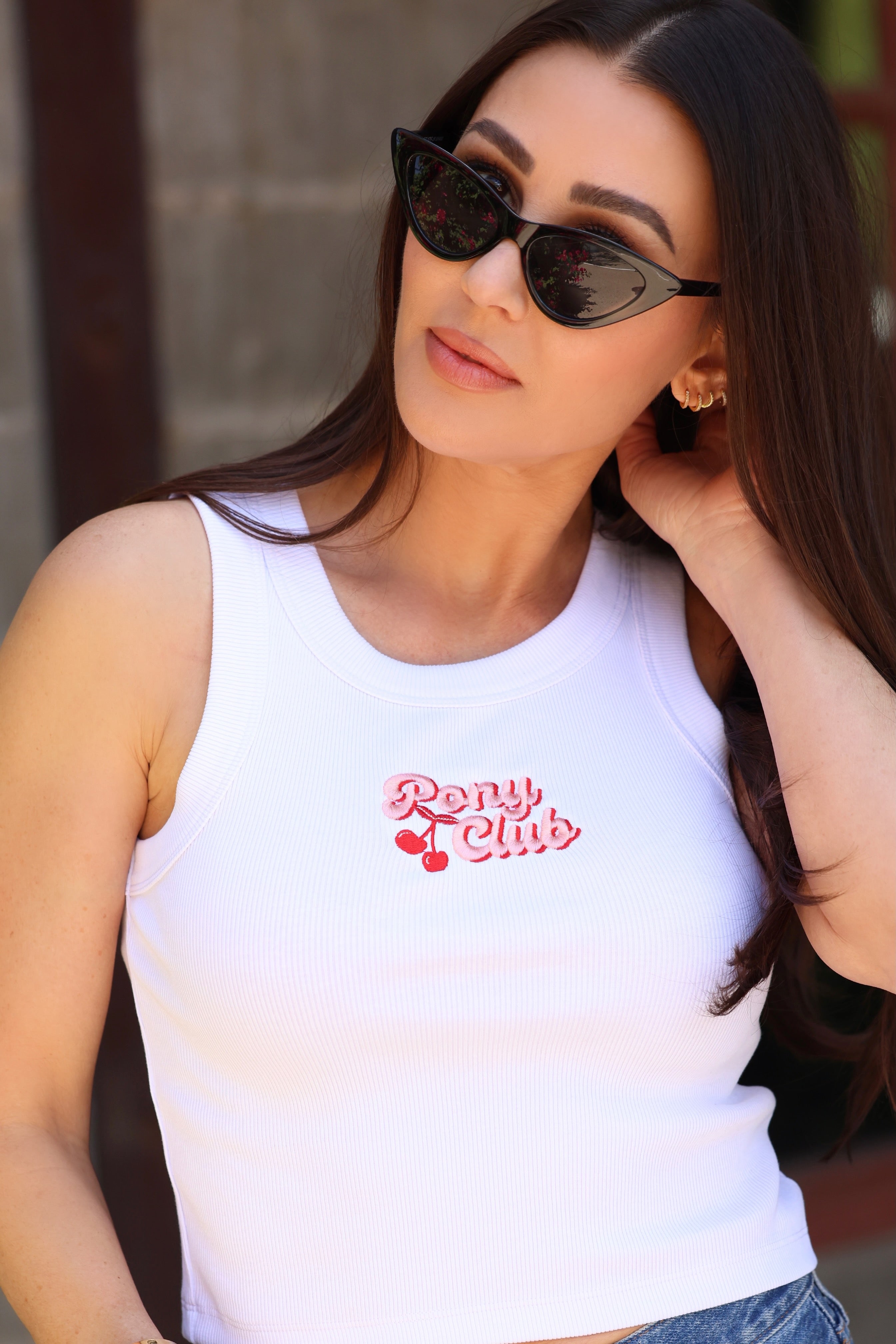 Woman in cat-eye sunglasses and white tank top with "Pony Club" logo, featuring cherries. She has long hair, a thoughtful expression, and touches her neck.