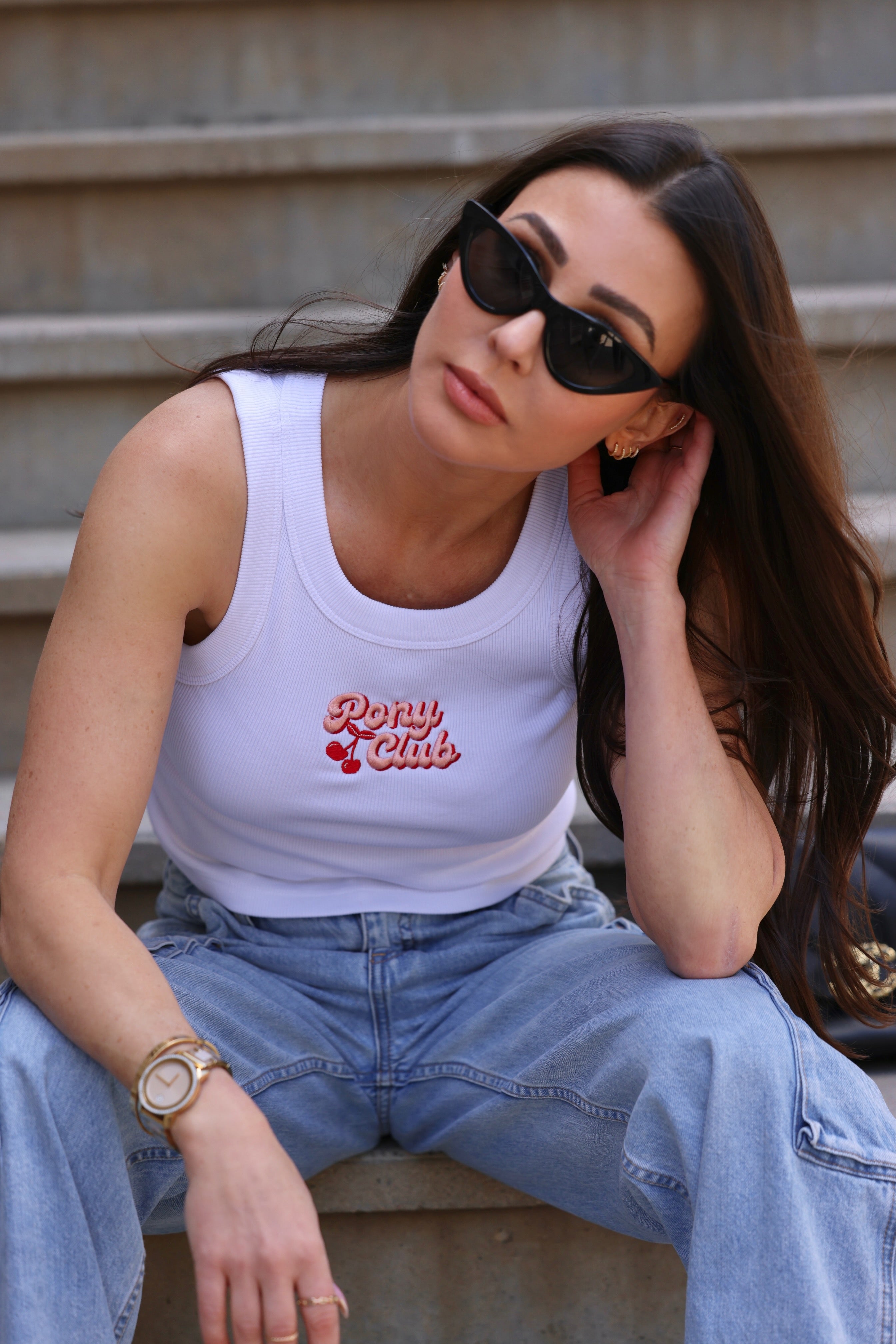 Woman with long brown hair sits on steps, wearing black sunglasses, a white tank top with "Pony Club," and blue jeans. Casual and relaxed mood.