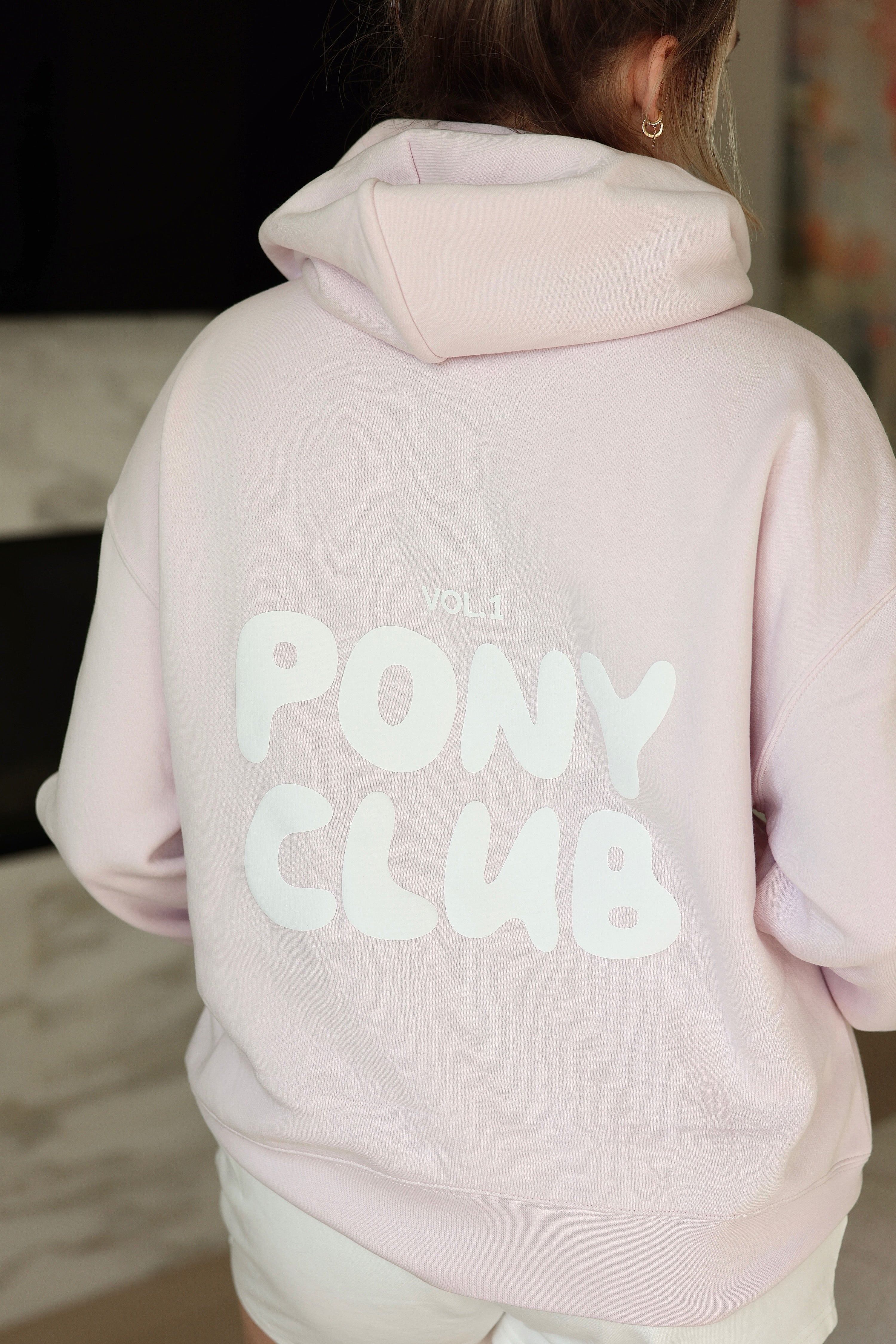 Rear view of a person wearing a pastel petal pink hoodie with "Vol 1 PONY CLUB" printed in bold white letters. The setting is casual and relaxed.