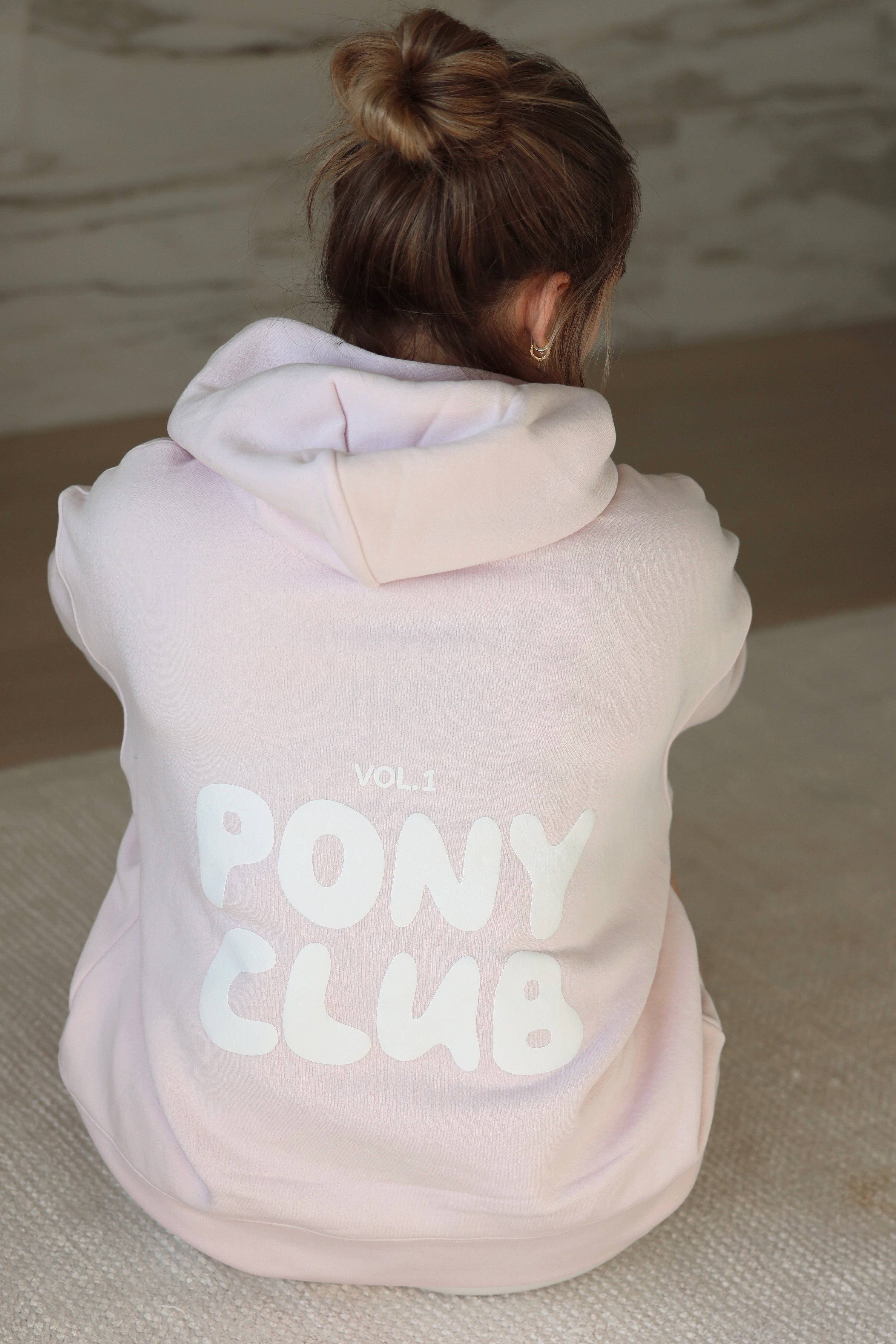 Person wearing a petal pink hoodie with "Pony Club Vol. 1" printed on the back. Their hair is in a messy bun, sitting on a textured carpet, conveying a cozy vibe.