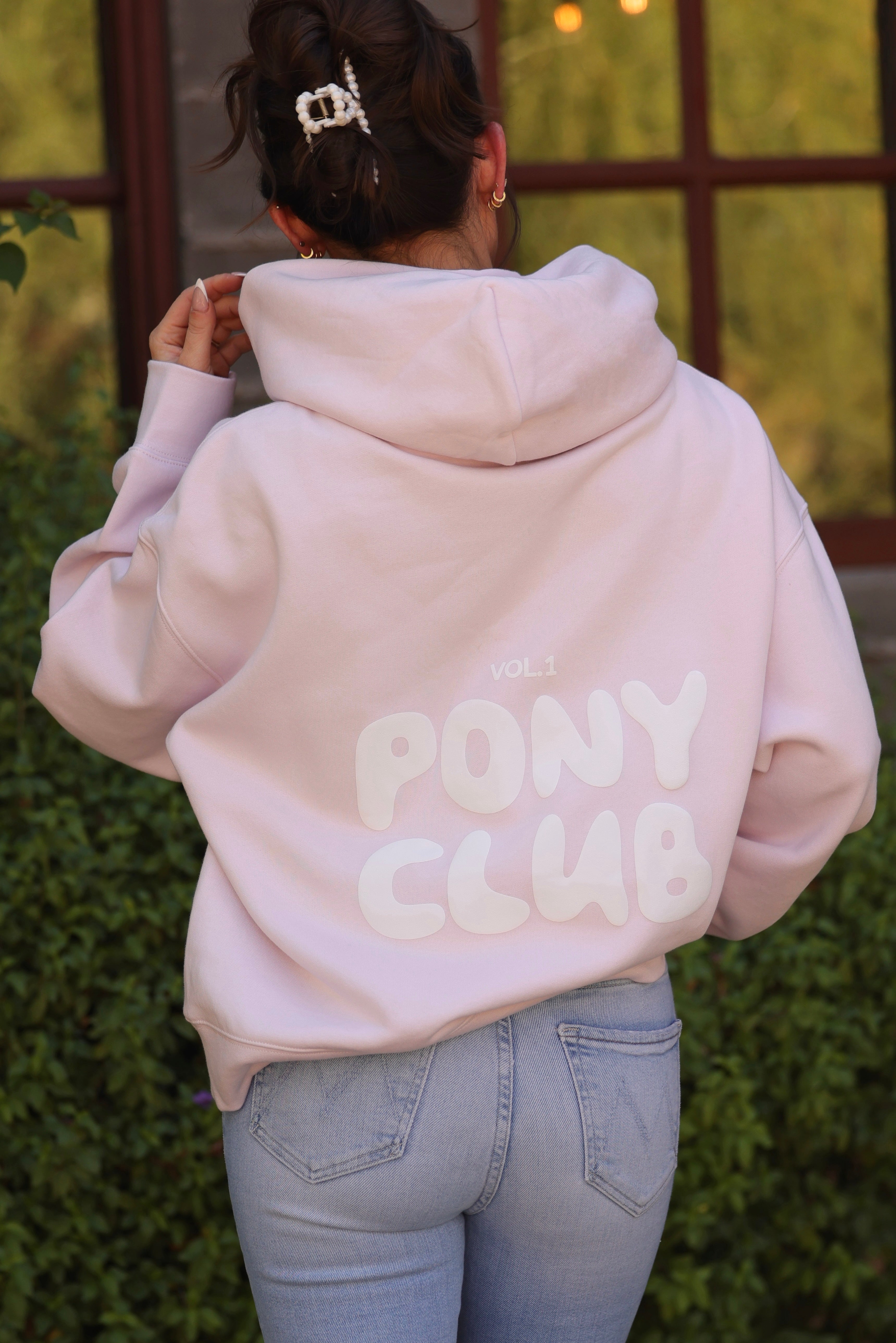 Rear view of a person wearing a pastel petal pink hoodie with "Vol 1 PONY CLUB" printed in bold white letters. The setting is casual and relaxed.