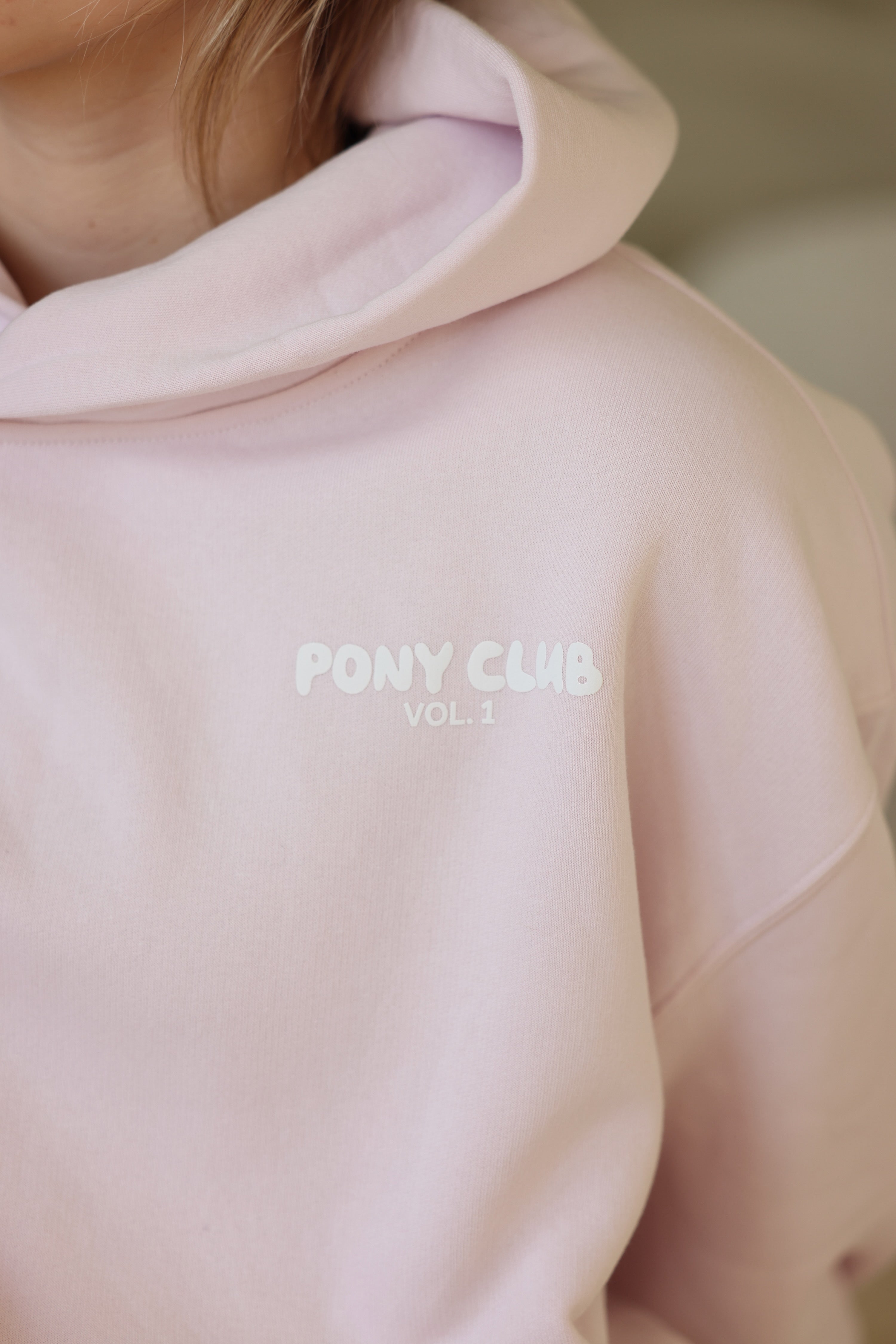 Close-up of a person wearing a light petal pink hoodie with "PONY CLUB VOL. 1" in white text. The fabric appears soft, conveying a cozy feel.