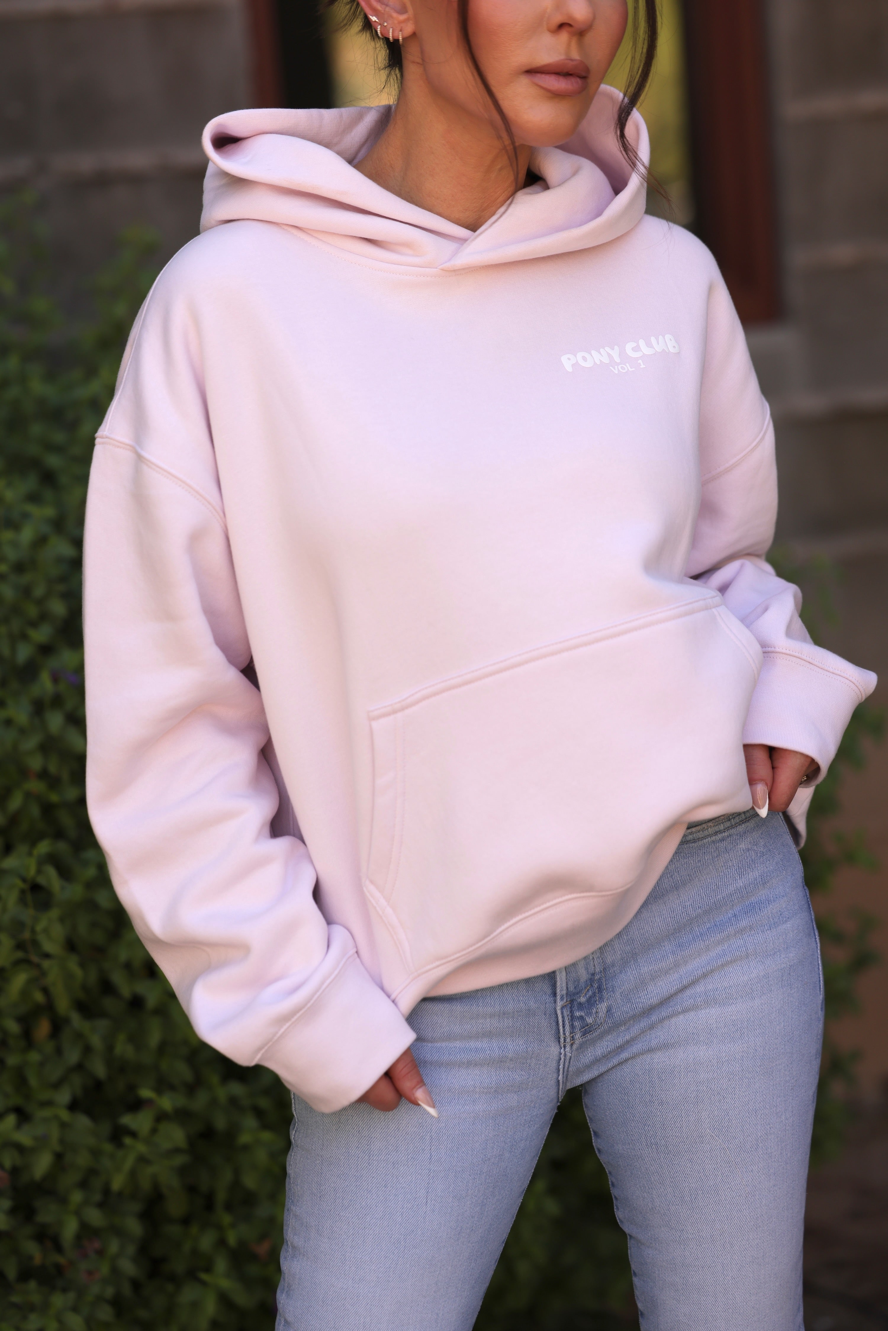 Close-up of a person wearing a light petal pink hoodie with "PONY CLUB VOL. 1" in white text. The fabric appears soft, conveying a cozy feel.