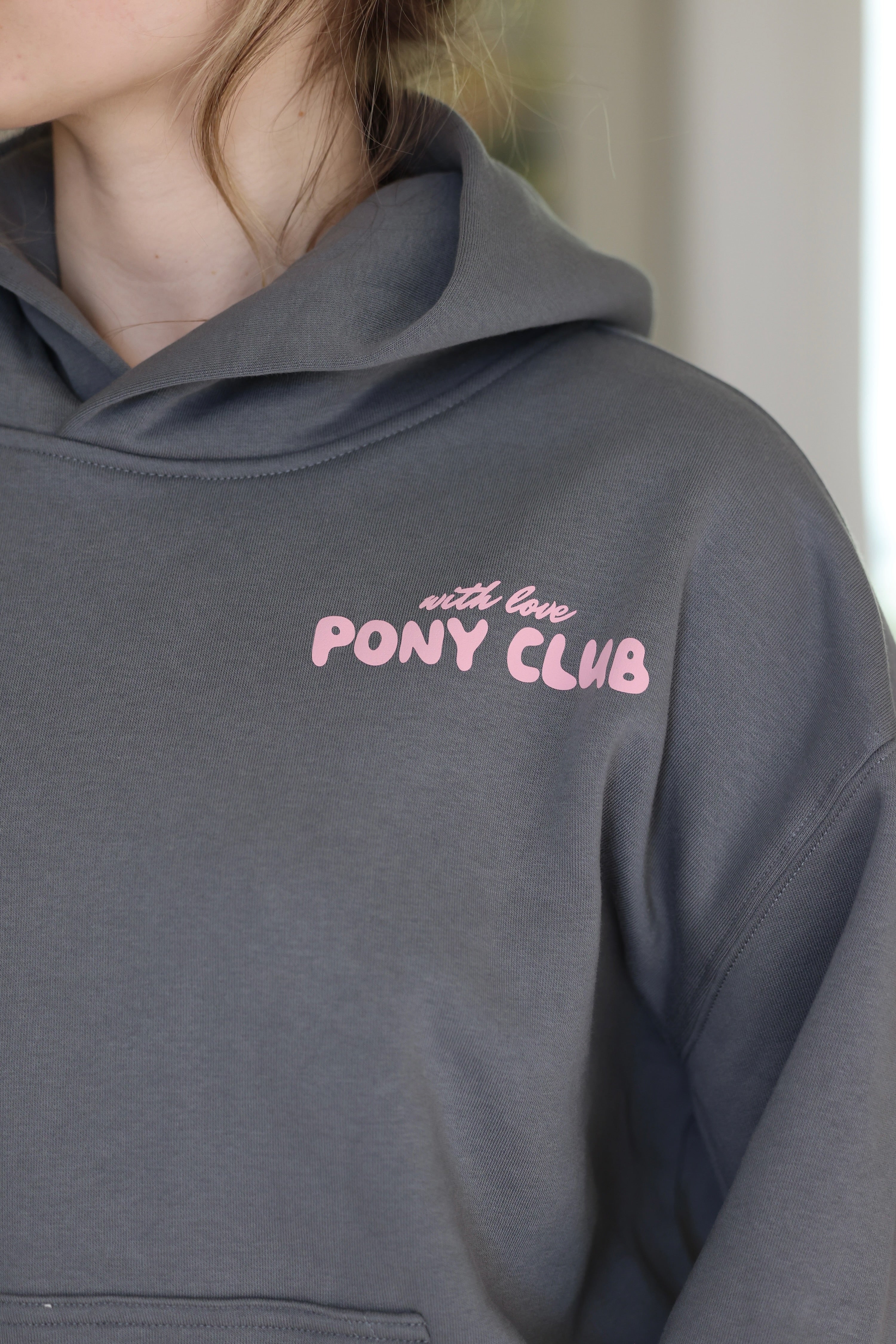 Close-up of a shadow gray hoodie with "with love PONY CLUB" printed in pink on the chest. The image conveys a casual and cozy feel.