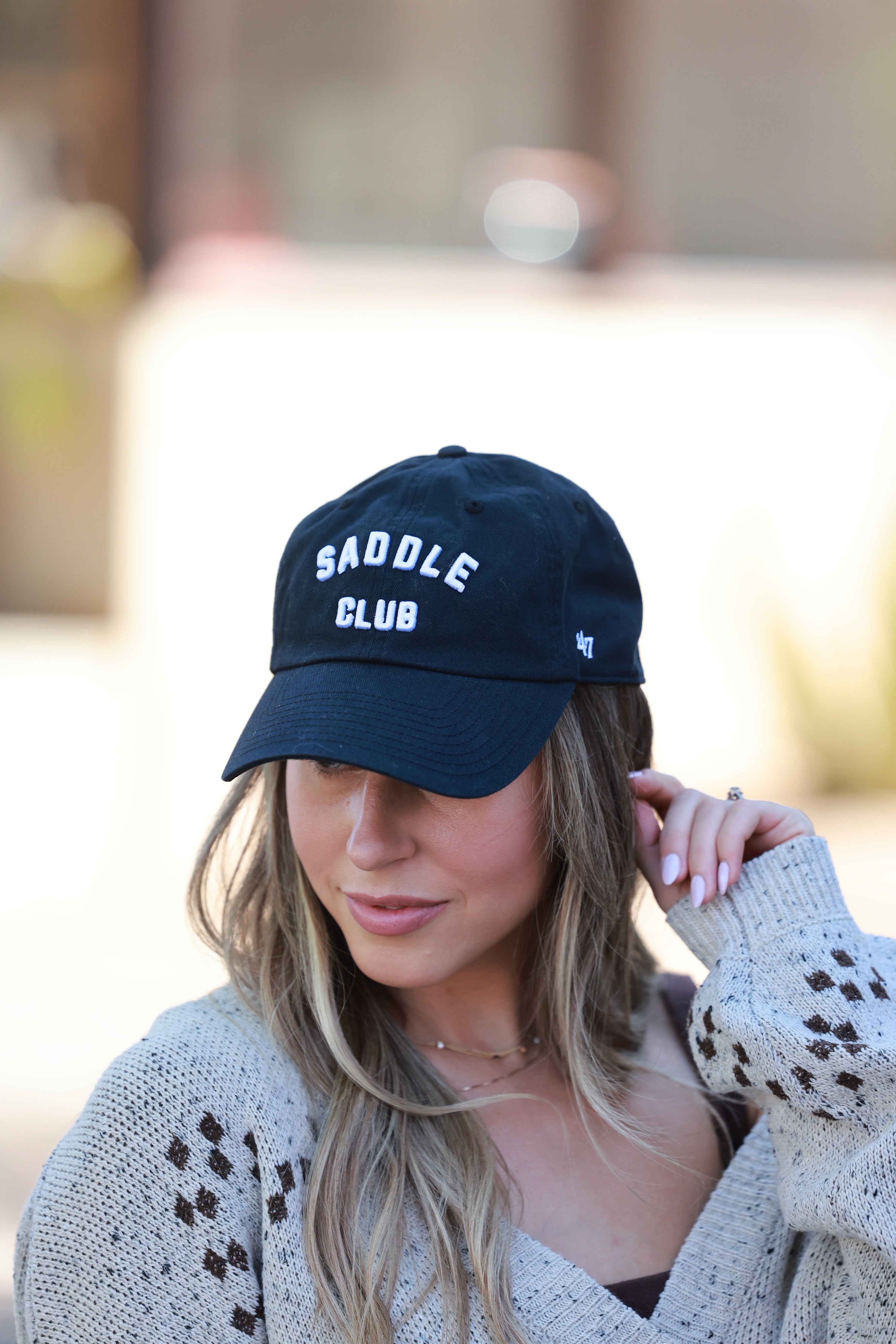 Woman wearing a black cap with 'Saddle Club' text, blurred background - equestrian accessory, horse riding gear, horse girl baseball cap, horse show hat