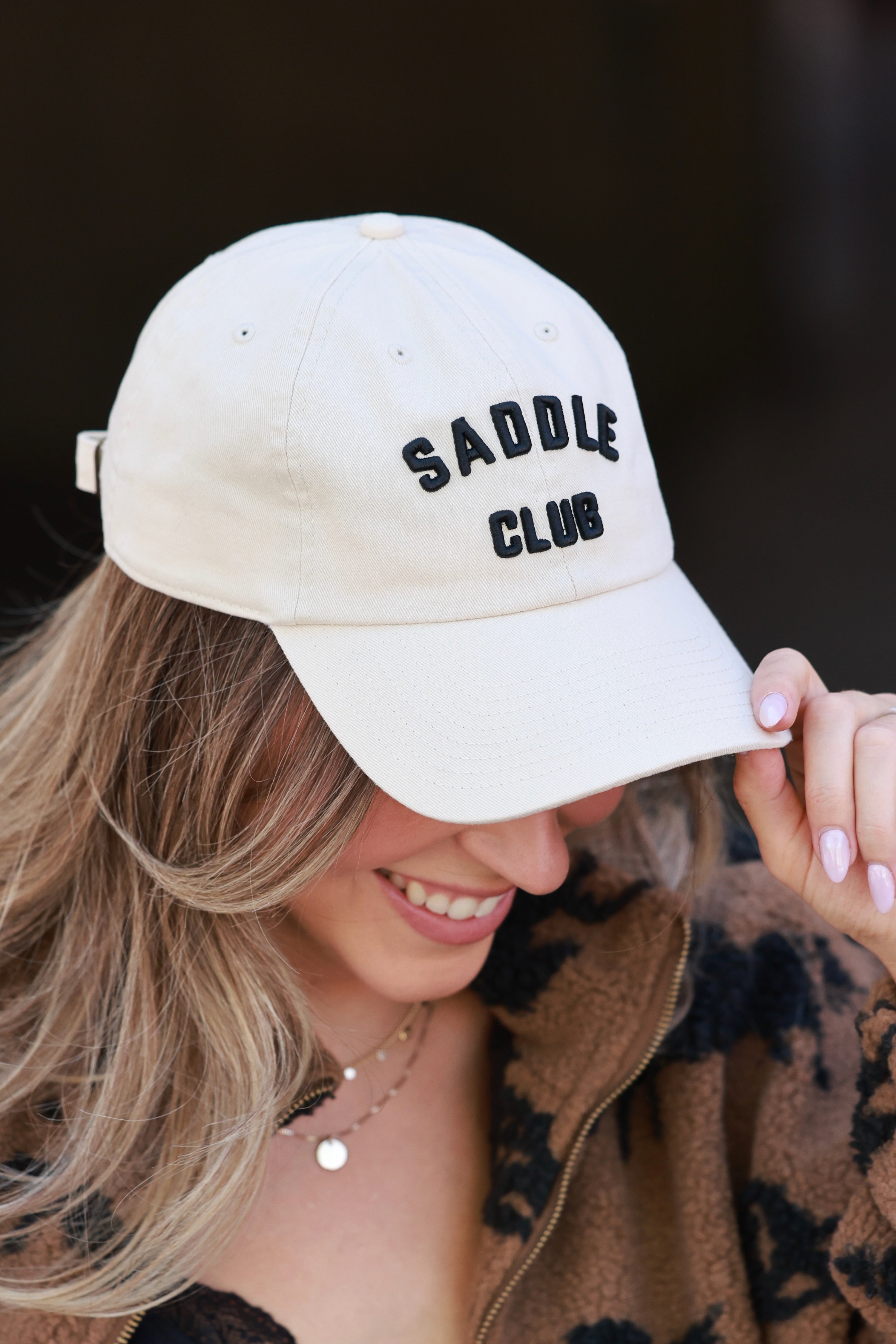 Person wearing a beige cap with 'SADDLE CLUB' text against a dark - equestrian accessory, horse riding gear, horse girl baseball cap, horse show hatbackground