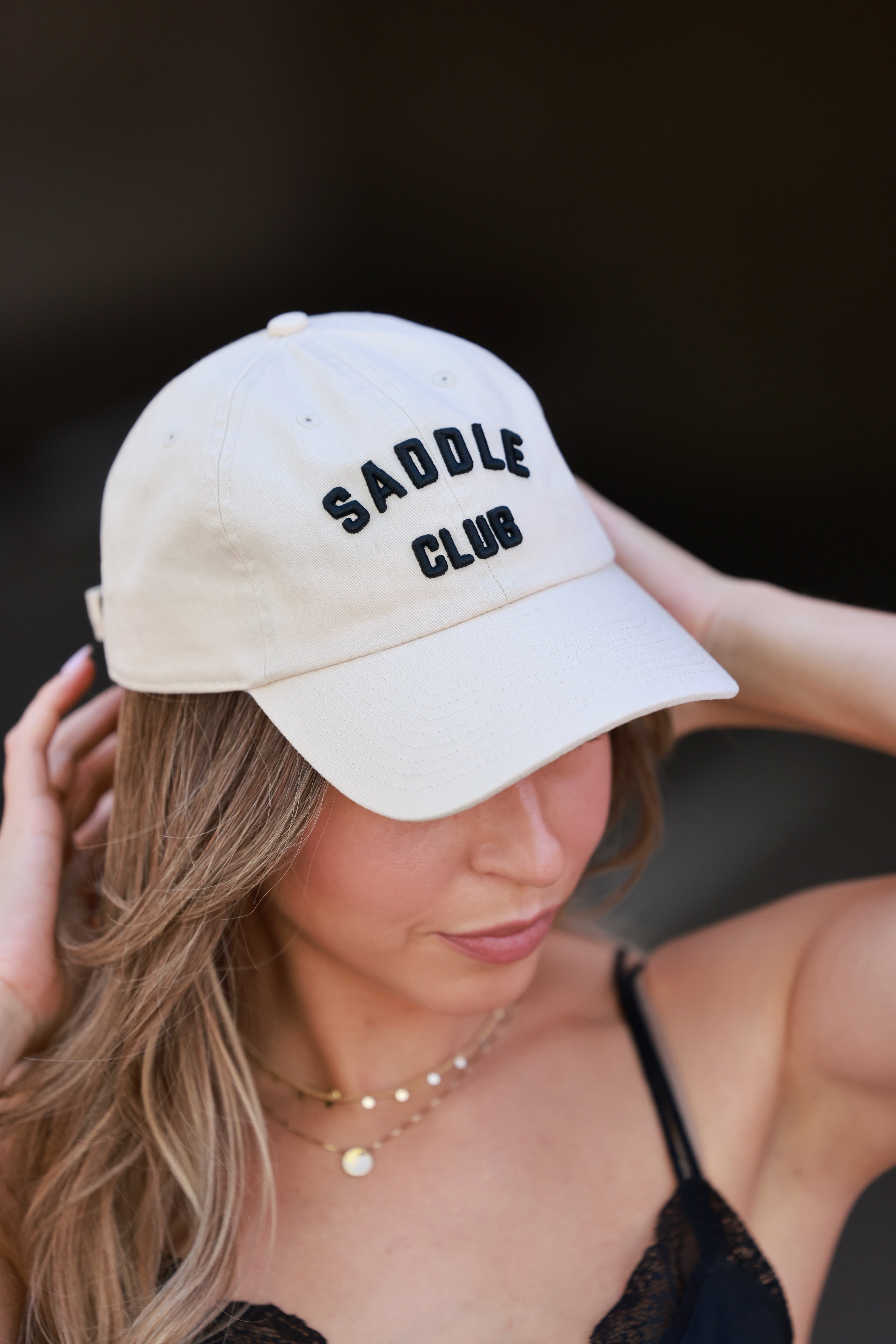 Woman wearing a beige cap with 'SADDLE CLUB' text against a dark - equestrian accessory, horse riding gear, horse girl baseball cap, horse show hatbackground