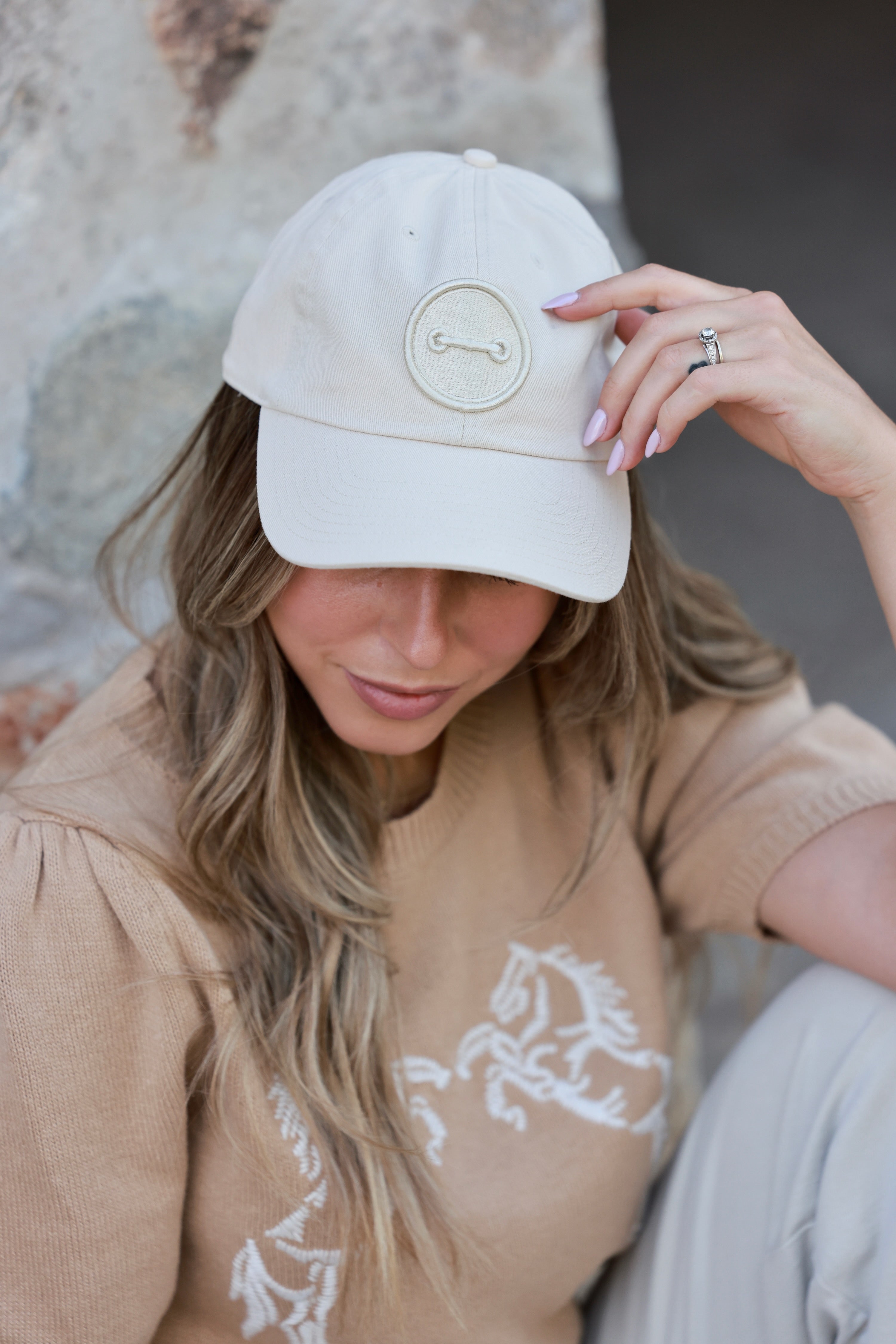 Woman wearing a beige cap with a logo, sitting outdoors. - horse bit hat, equestrian headwear, horse girl accessories, casual riding gear, horse riding accessories, baseball cap