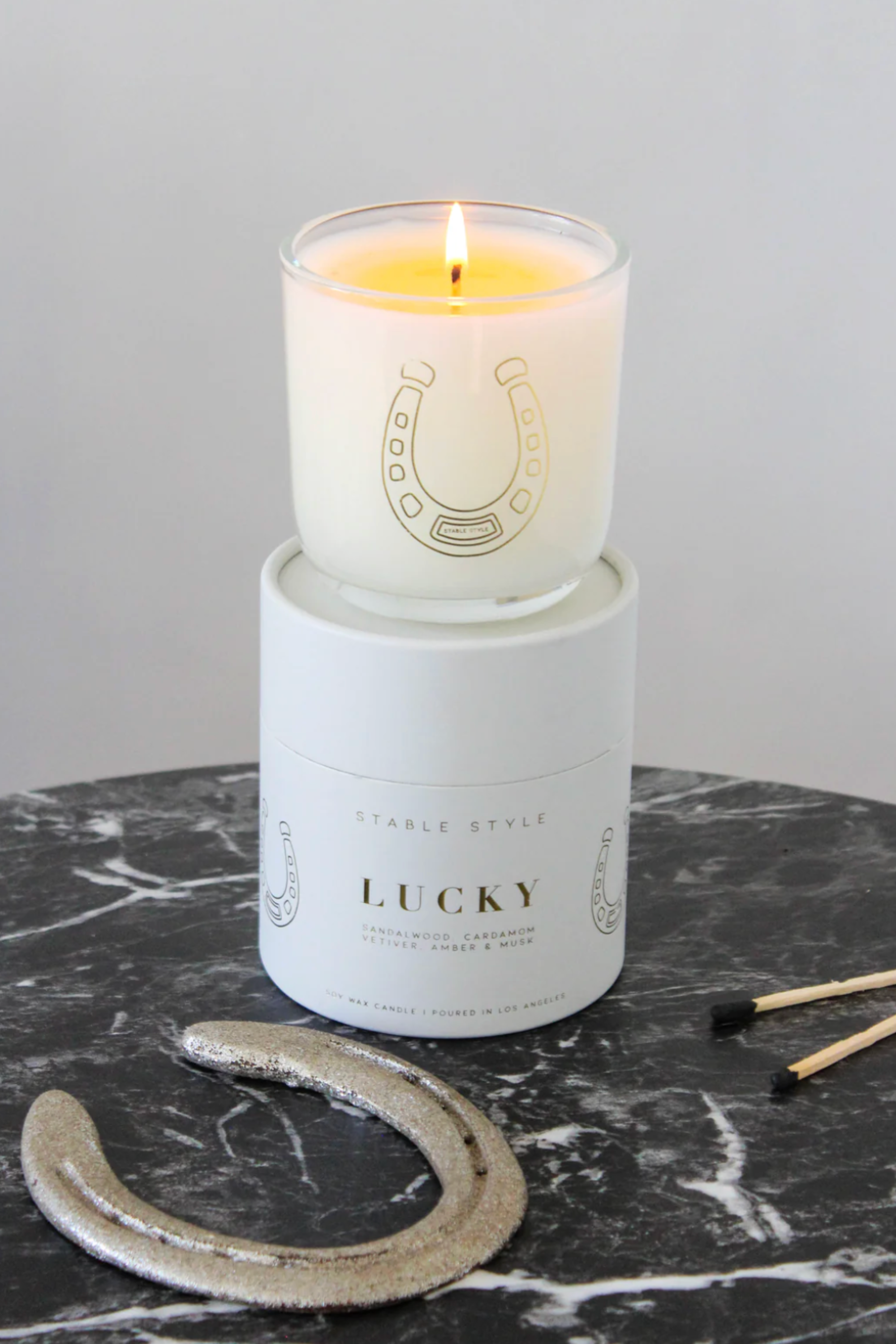 Stable Style Icons Collection 'Lucky Horseshoe' candle — a 10-ounce soy wax candle hand-poured into a clear glass vessel with a rounded base and gold accents. The fragrance opens with bright lemon and spicy cardamom, followed by a warm cedarwood–sandalwood heart with cinnamon and nutmeg, and finishes with earthy vetiver, amber, and musk. Presented in a gold-foil wrapped paper tube, offering 60+ hours of burn time.