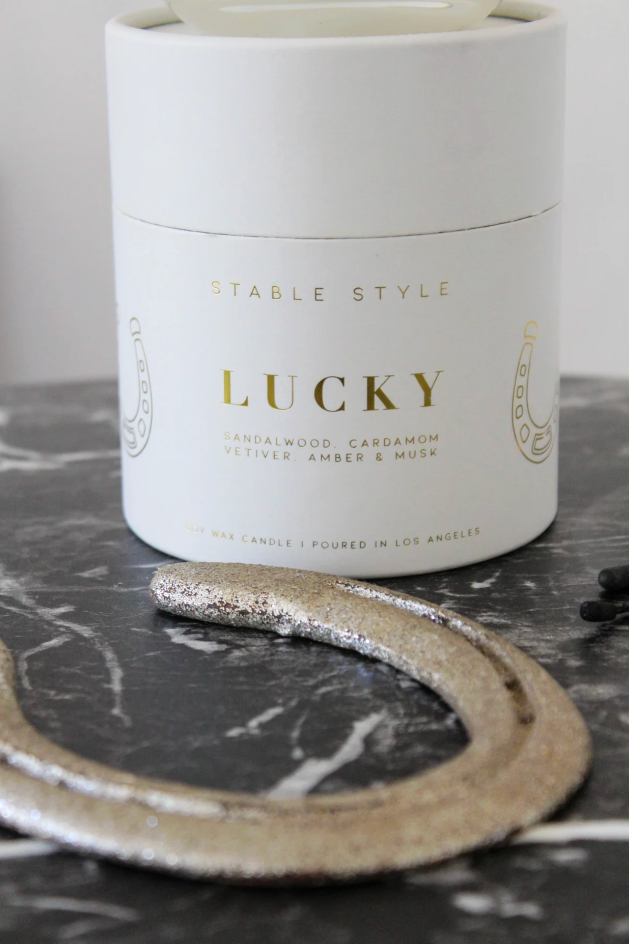 Stable Style Icons Collection 'Lucky Horseshoe' candle — a 10-ounce soy wax candle hand-poured into a clear glass vessel with a rounded base and gold accents. The fragrance opens with bright lemon and spicy cardamom, followed by a warm cedarwood–sandalwood heart with cinnamon and nutmeg, and finishes with earthy vetiver, amber, and musk. Presented in a gold-foil wrapped paper tube, offering 60+ hours of burn time.