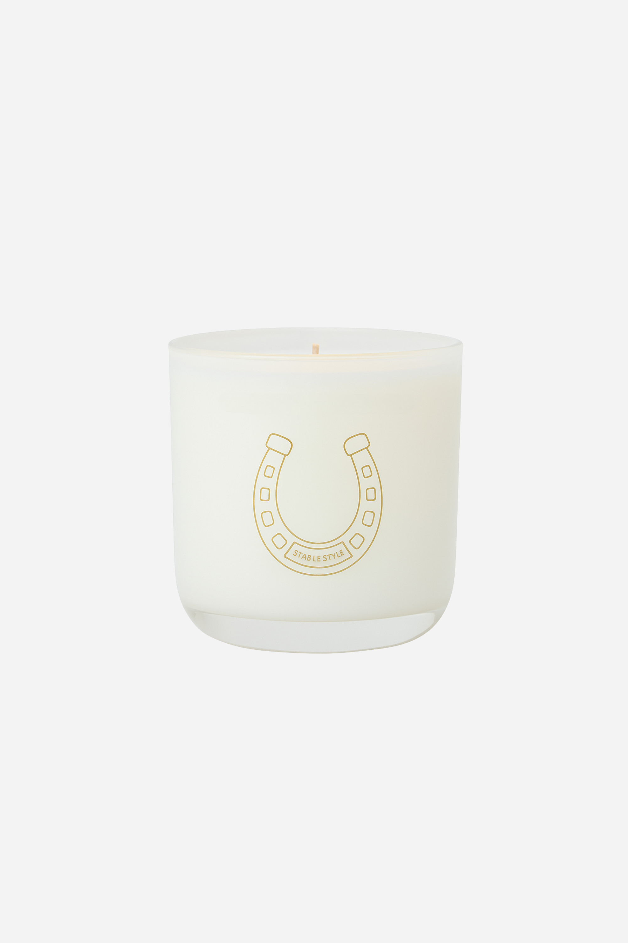 Stable Style Icons Collection 'Lucky Horseshoe' candle — a 10-ounce soy wax candle hand-poured into a clear glass vessel with a rounded base and gold accents. The fragrance opens with bright lemon and spicy cardamom, followed by a warm cedarwood–sandalwood heart with cinnamon and nutmeg, and finishes with earthy vetiver, amber, and musk. Presented in a gold-foil wrapped paper tube, offering 60+ hours of burn time.