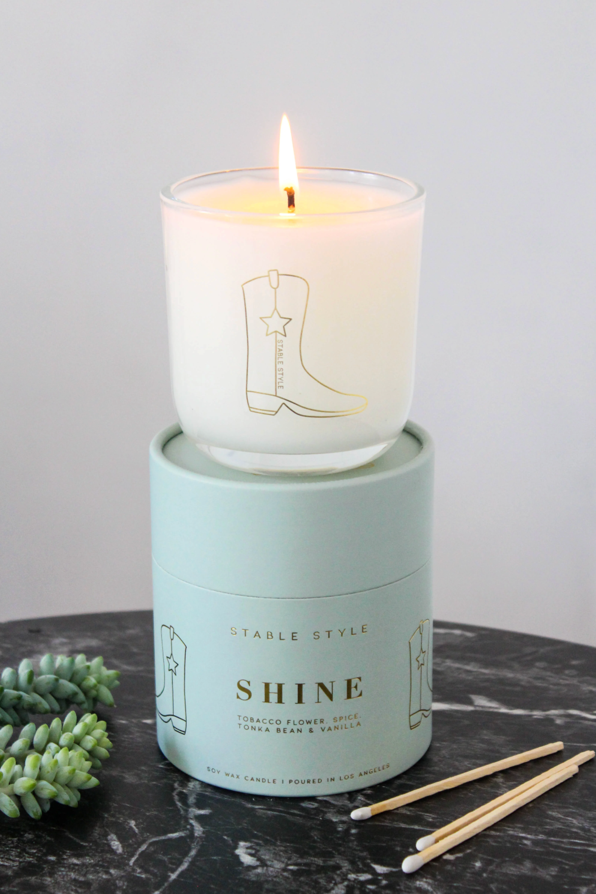 10-ounce soy wax candle from the Icons Collection by Stable Style, hand-poured into a clear glass vessel with a rounded base and gold accents. Fragrance opens with tobacco flower, anise, nutmeg, clove, and ginger, with warm mid notes of vanilla, tonka bean, and cocoa, and a rich base of amber, agarwood, and sweet fruit. Elegant packaging comes in a gold-foil wrapped paper tube, offering 60+ hours of burn time.