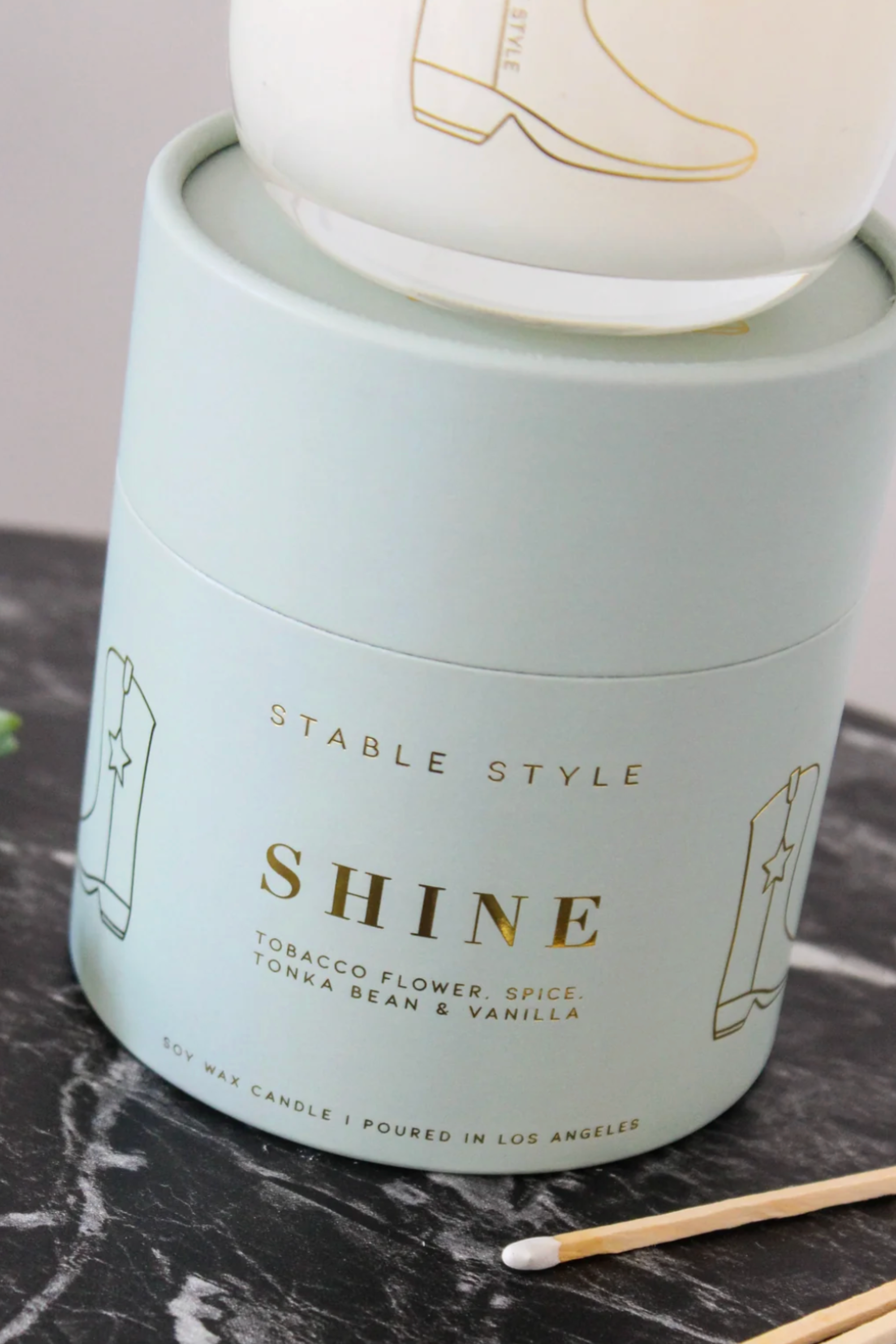 10-ounce soy wax candle from the Icons Collection by Stable Style, hand-poured into a clear glass vessel with a rounded base and gold accents. Fragrance opens with tobacco flower, anise, nutmeg, clove, and ginger, with warm mid notes of vanilla, tonka bean, and cocoa, and a rich base of amber, agarwood, and sweet fruit. Elegant packaging comes in a gold-foil wrapped paper tube, offering 60+ hours of burn time.
