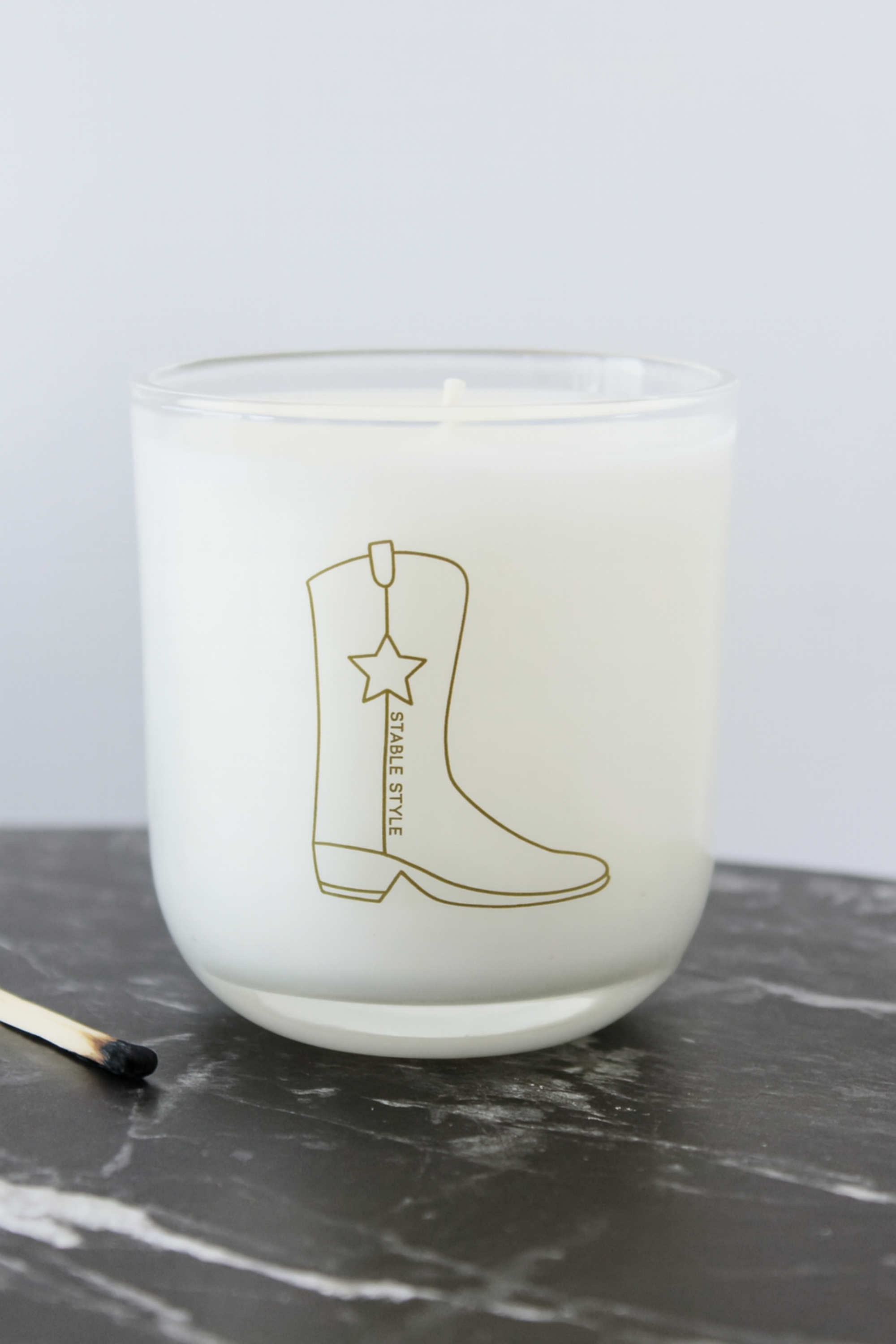 10-ounce soy wax candle from the Icons Collection by Stable Style, hand-poured into a clear glass vessel with a rounded base and gold accents. Fragrance opens with tobacco flower, anise, nutmeg, clove, and ginger, with warm mid notes of vanilla, tonka bean, and cocoa, and a rich base of amber, agarwood, and sweet fruit. Elegant packaging comes in a gold-foil wrapped paper tube, offering 60+ hours of burn time.