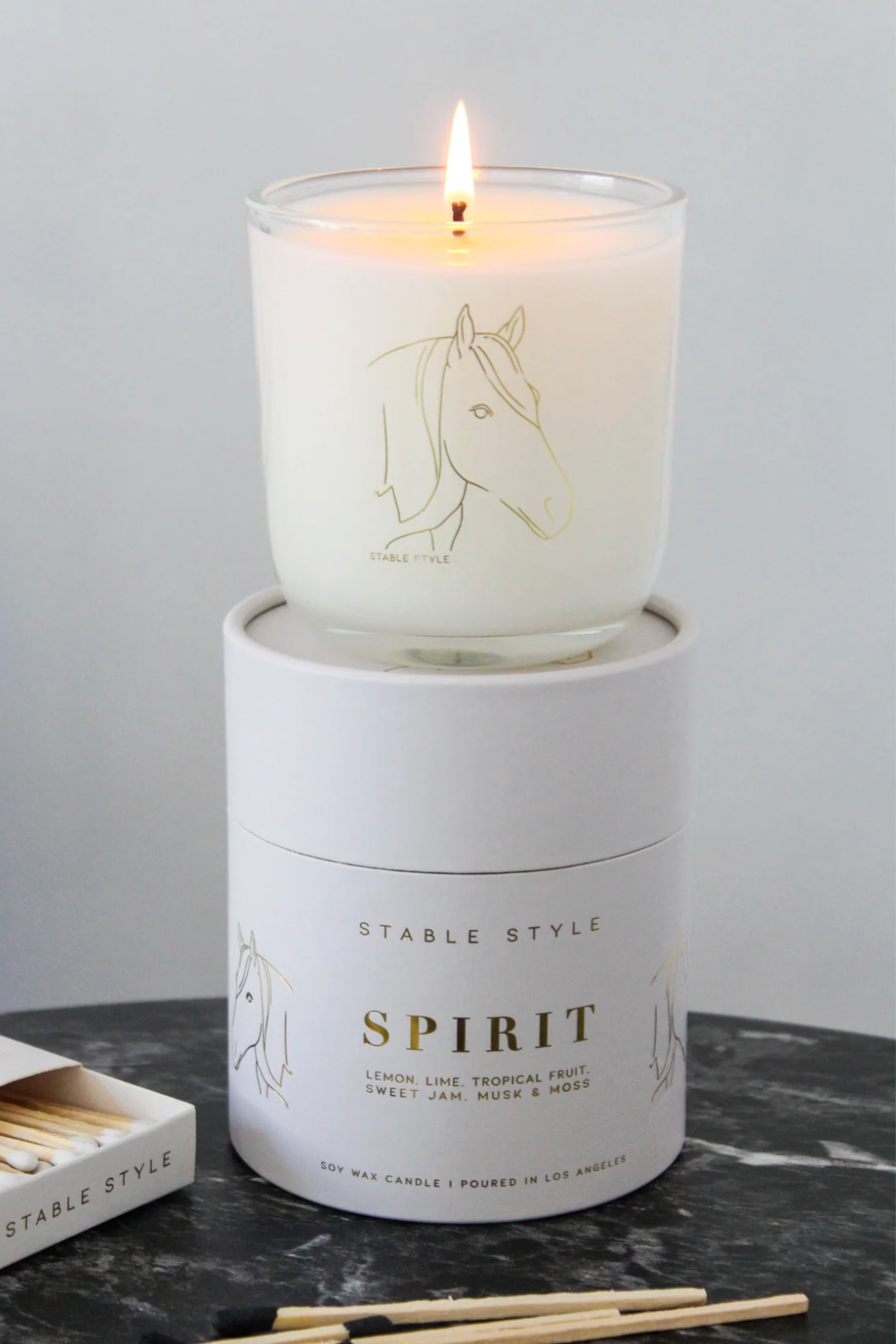White soy wax candle labeled 'Spirit,' featuring elegant horse illustrations, set on marble with matchsticks nearby. Fragrance opens with bright lemon and lime, transitions into tropical fruit and Tahitian gardenia, and finishes with sweet jam and warm musk. Hand-poured in a 10-ounce clear glass vessel with gold accents, offering 60+ hours of burn time.