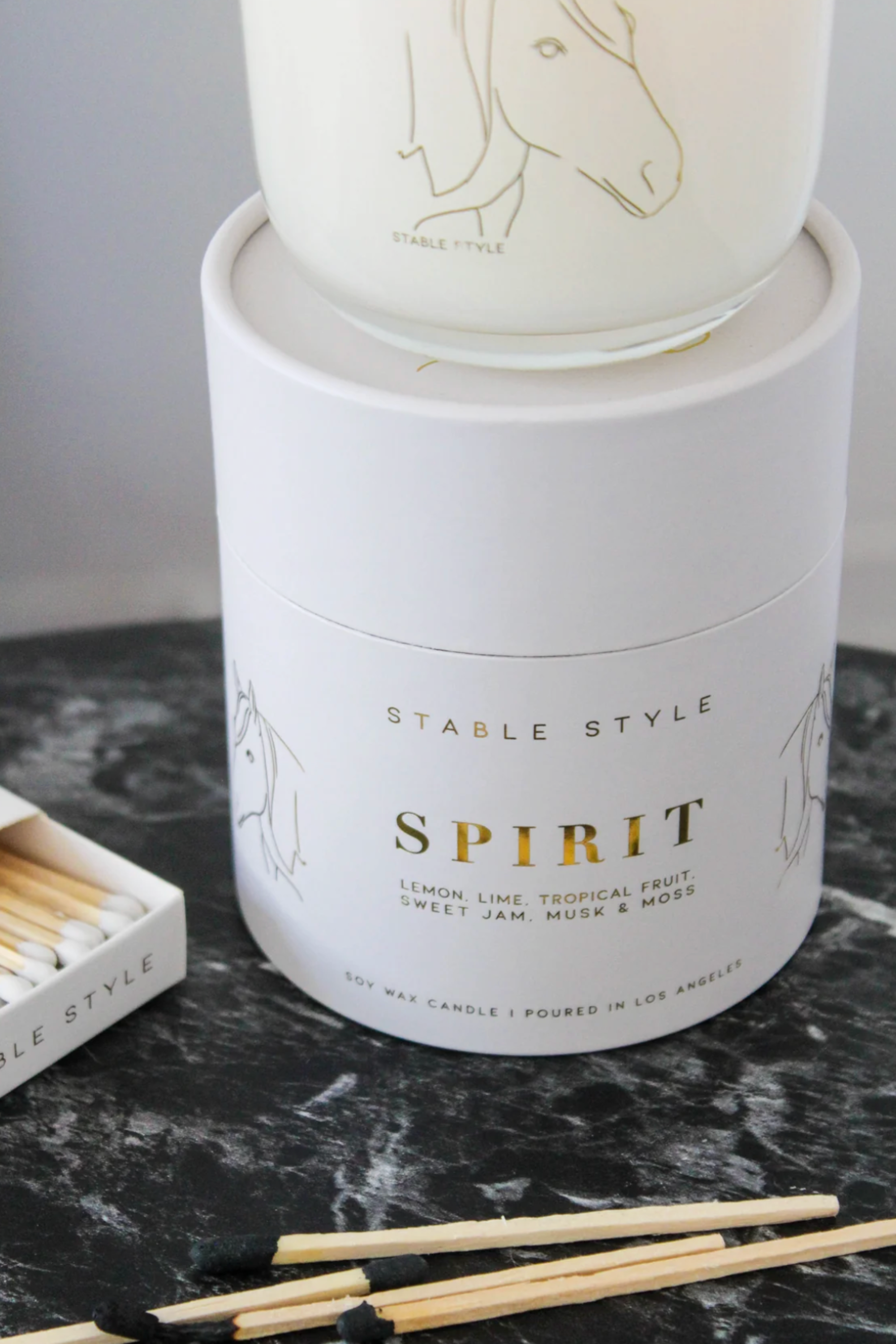 White soy wax candle labeled 'Spirit,' featuring elegant horse illustrations, set on marble with matchsticks nearby. Fragrance opens with bright lemon and lime, transitions into tropical fruit and Tahitian gardenia, and finishes with sweet jam and warm musk. Hand-poured in a 10-ounce clear glass vessel with gold accents, offering 60+ hours of burn time.