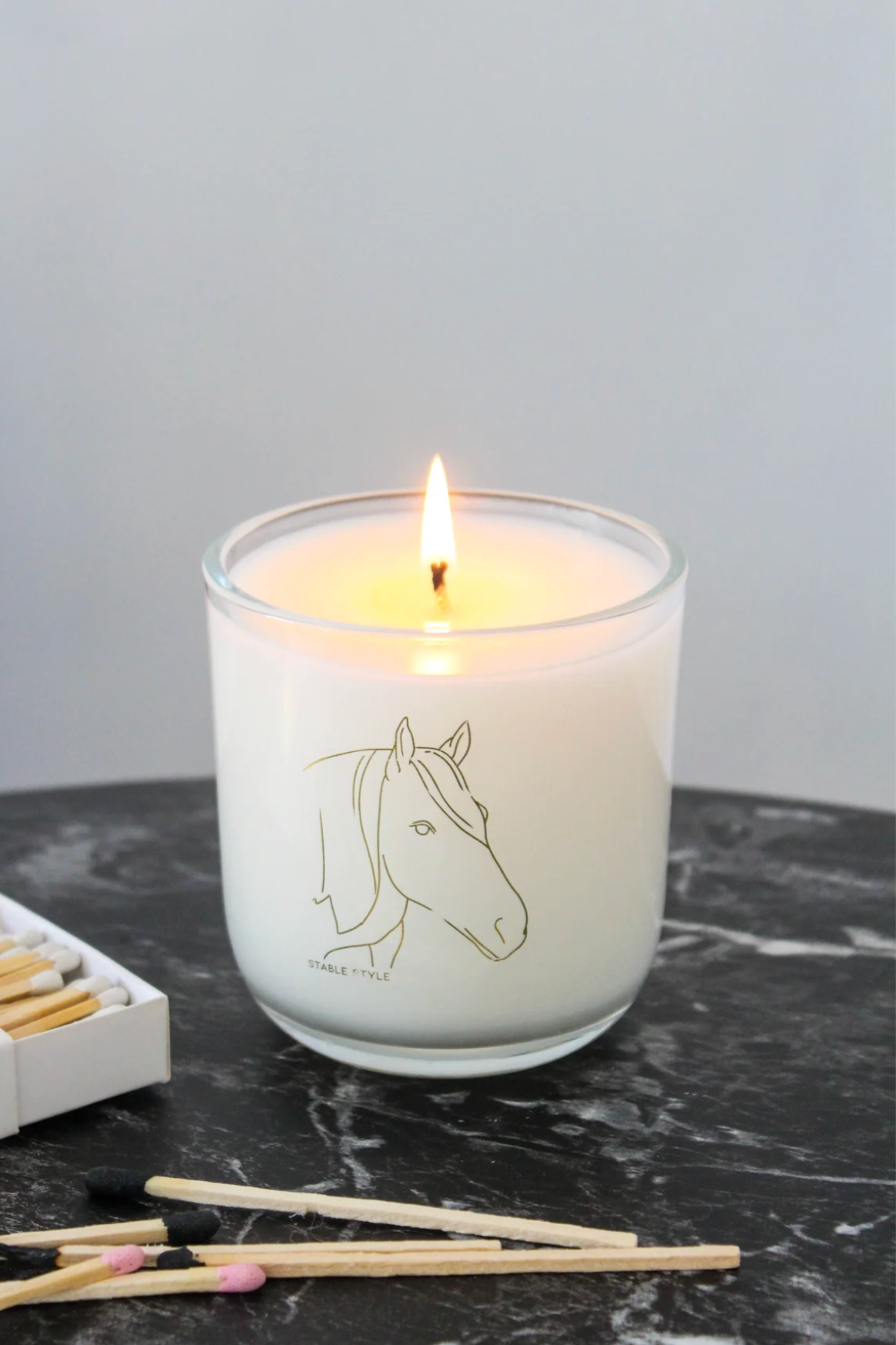White soy wax candle labeled 'Spirit,' featuring elegant horse illustrations, set on marble with matchsticks nearby. Fragrance opens with bright lemon and lime, transitions into tropical fruit and Tahitian gardenia, and finishes with sweet jam and warm musk. Hand-poured in a 10-ounce clear glass vessel with gold accents, offering 60+ hours of burn time.