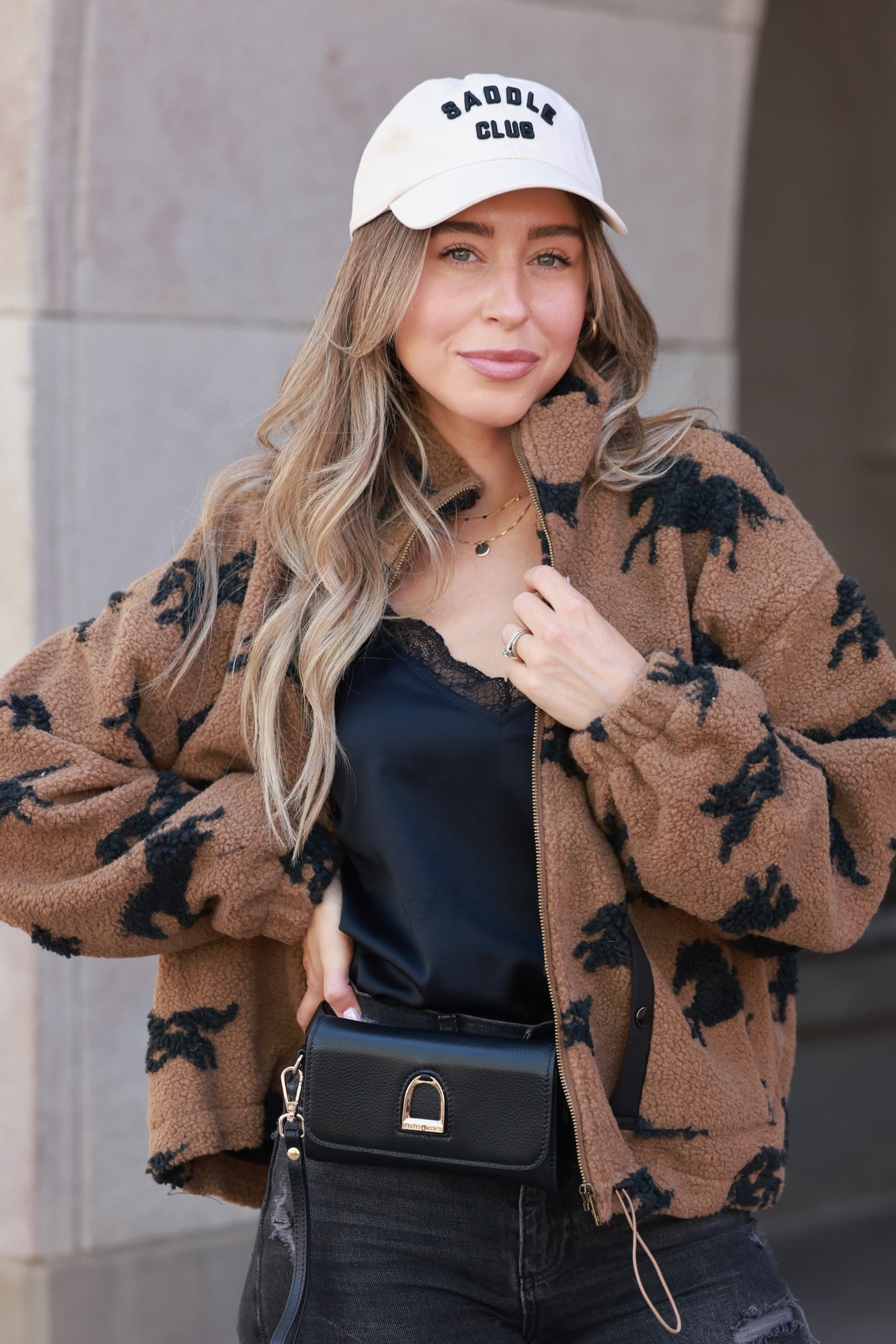 Woman wearing a brown patterned jacket, black top, and ripped jeans standing against a building. - equestrian sherpa jacket, equestrian jacket, horse print jacket, equestrian outerwear