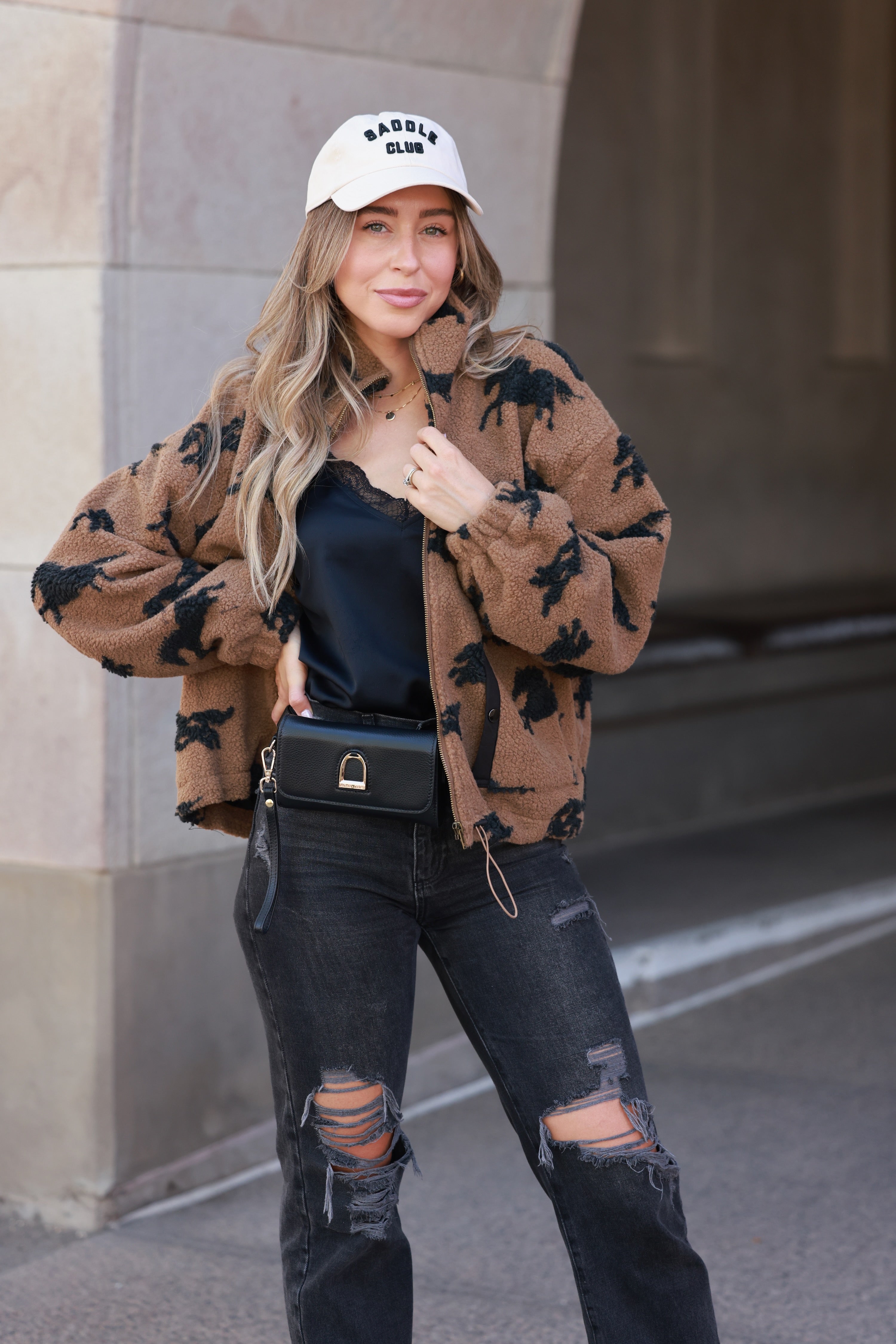 Woman wearing a brown patterned jacket, black top, and ripped jeans standing against a building. - equestrian sherpa jacket, equestrian jacket, horse print jacket, equestrian outerwear